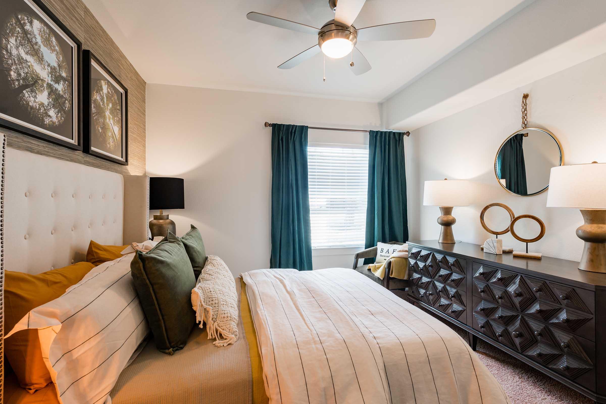 A stylish bedroom featuring a light beige and green color palette. The room includes a large upholstered bed with decorative pillows, a dark wooden dresser with circular mirrors, and stylish lamps. Sunlight filters through sheer curtains, creating a warm and inviting atmosphere. A ceiling fan adds a modern touch.