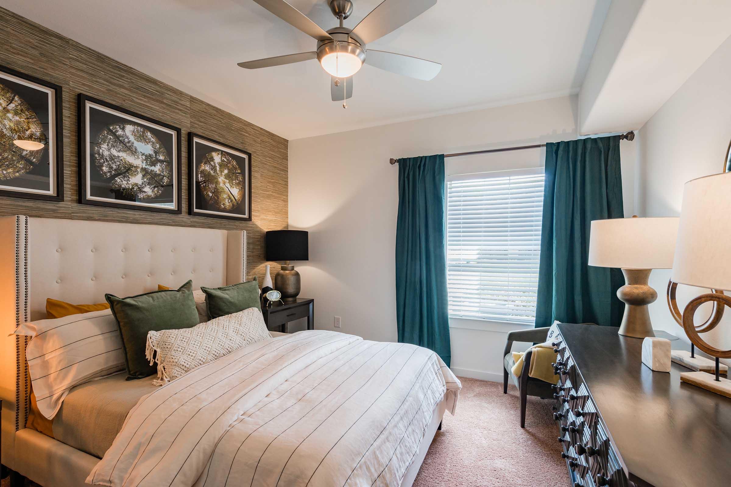 Cozy bedroom featuring a queen-sized bed with striped white and beige bedding, accented with green pillows. The room has a stylish tufted headboard, a wooden nightstand with a lamp, a decorative chest, and framed artworks on the wall. Natural light flows through a window with teal curtains.