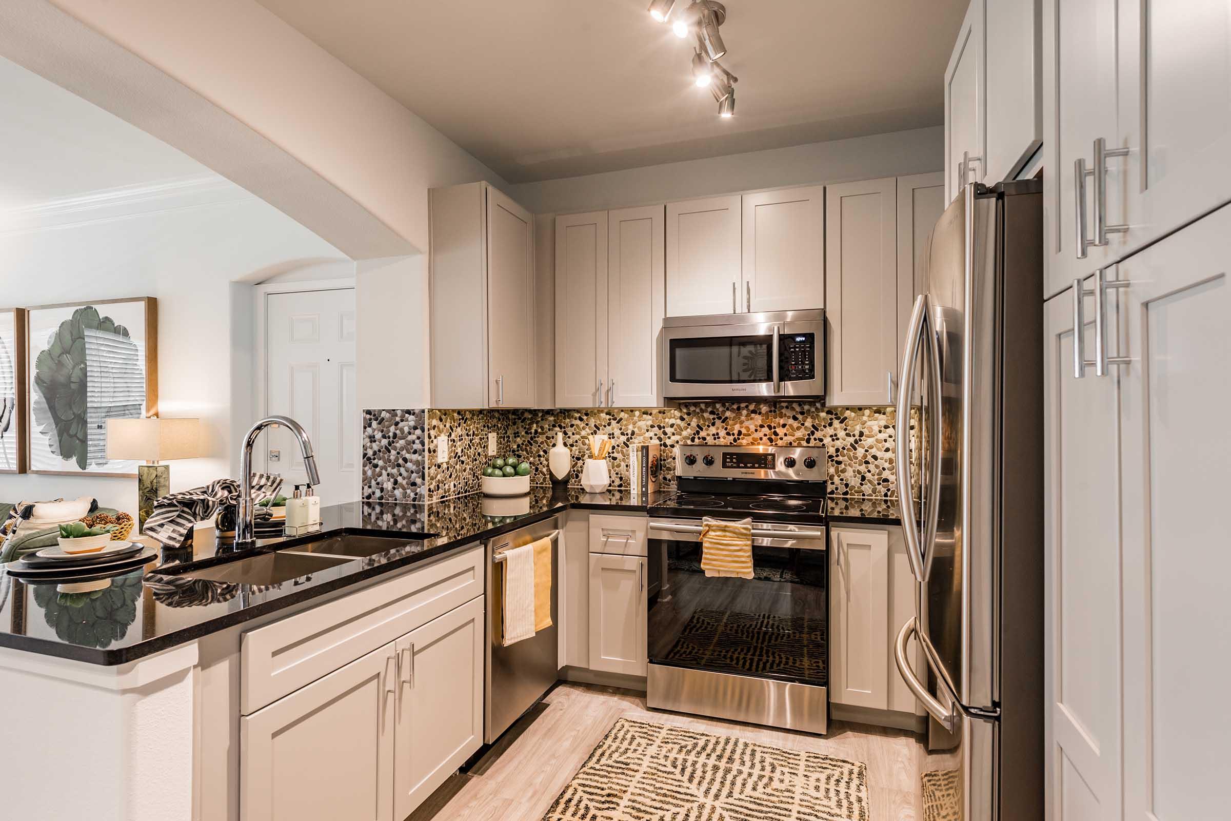 Modern kitchen featuring sleek gray cabinetry, stainless steel appliances including a microwave and oven, a black countertop, and a colorful mosaic backsplash. The space includes a double basin sink and is accented by decorative items on the countertop. Natural light enhances the inviting atmosphere.