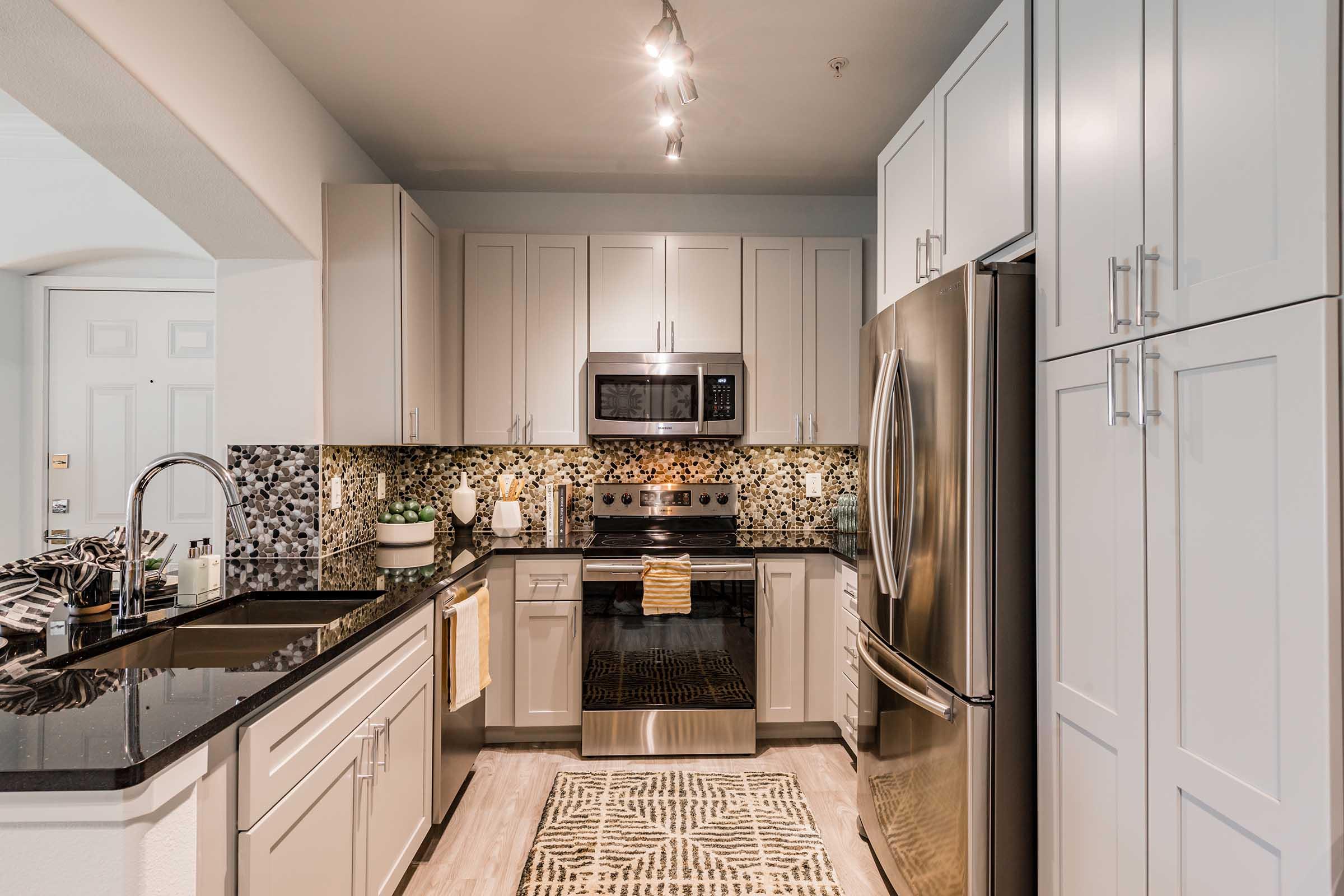 Modern kitchen featuring stainless steel appliances, including a microwave and a refrigerator. The counters are black granite, and the cabinetry is light gray. A mosaic tile backsplash adds texture, while a rug lies on the floor. Soft lighting highlights the space, creating an inviting atmosphere.