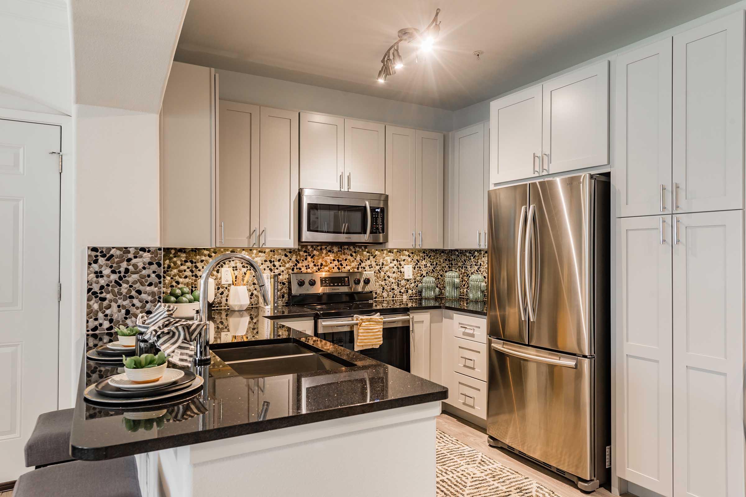 Modern kitchen featuring stainless steel appliances, including a microwave and refrigerator. The kitchen has sleek white cabinets, a dark granite countertop, and a stylish backsplash with a mosaic design. There are decorative items on the counter and a dining area with two stools. Brightly lit with recessed lighting.