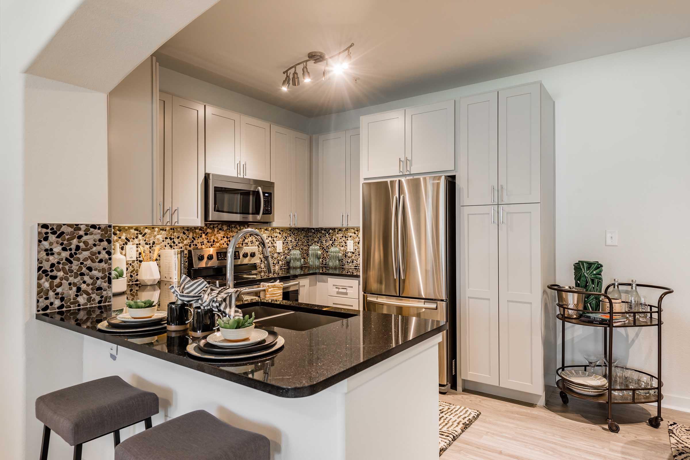 Modern kitchen featuring sleek gray cabinets, stainless steel appliances, and a black countertop. A mosaic backsplash adds a stylish touch. The kitchen island includes bar stools and is set with plates and decorative items. Soft lighting enhances the inviting atmosphere, complemented by a nearby cart with glassware.