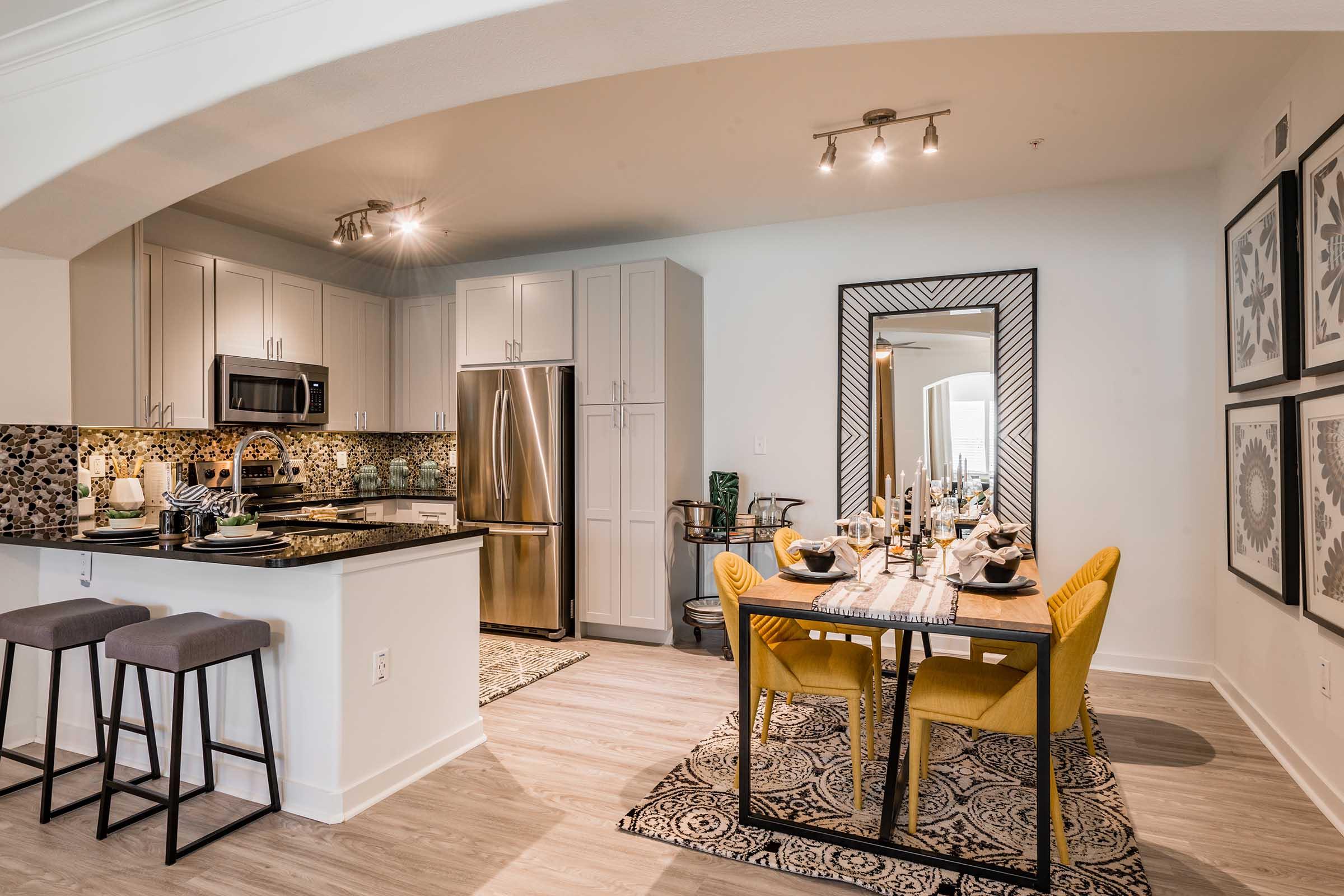 Modern kitchen and dining area featuring a stylish dining table with yellow chairs, black and stainless steel appliances, and a sleek countertop. The space includes decorative elements such as artwork on the walls and a patterned rug, creating an inviting atmosphere.