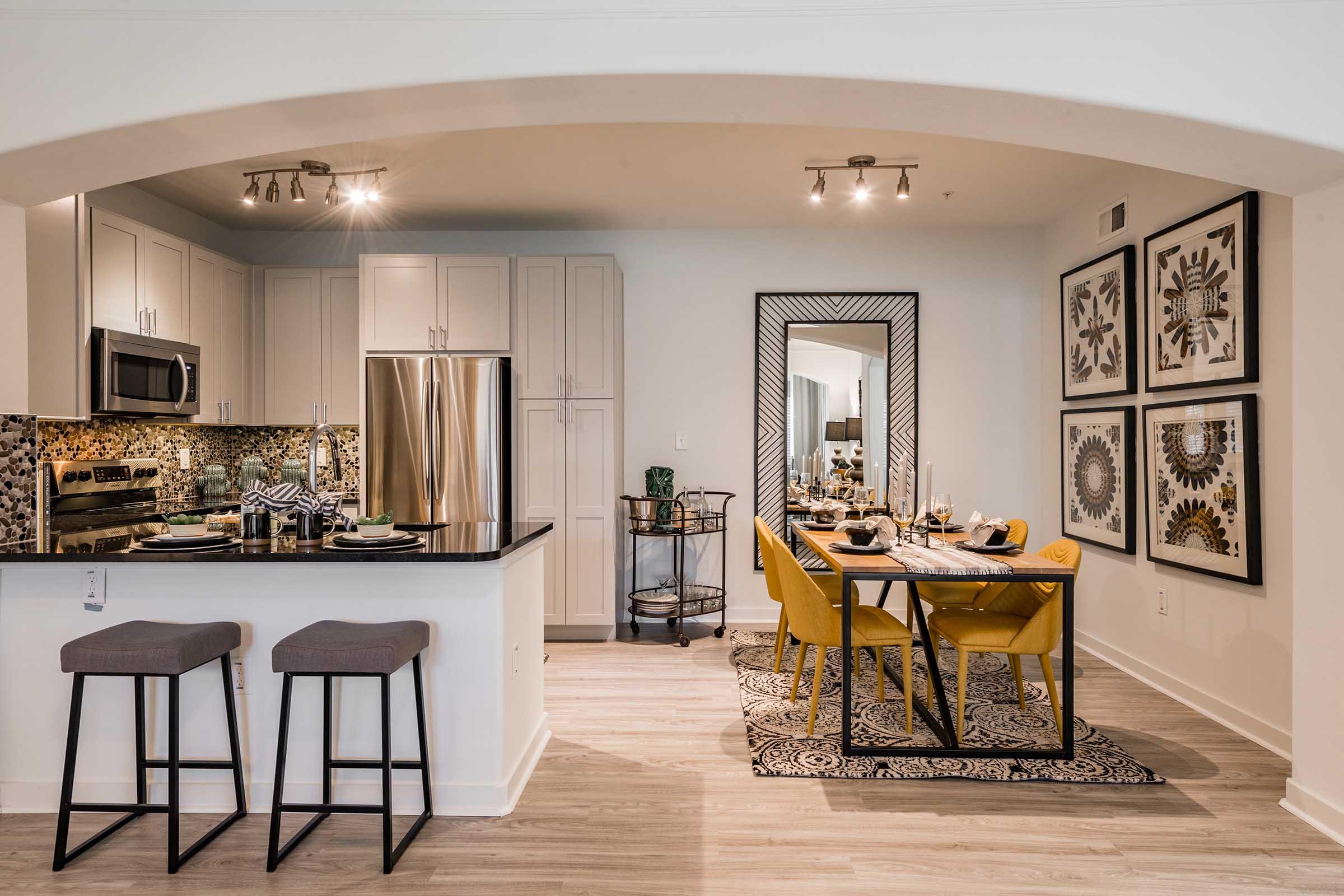 A modern kitchen and dining area featuring sleek cabinetry, stainless steel appliances, and a stylish dining table with yellow chairs. The space is illuminated by ceiling lights, and decorative artwork adorns the walls, creating an inviting atmosphere. Bar stools are positioned at the kitchen counter.