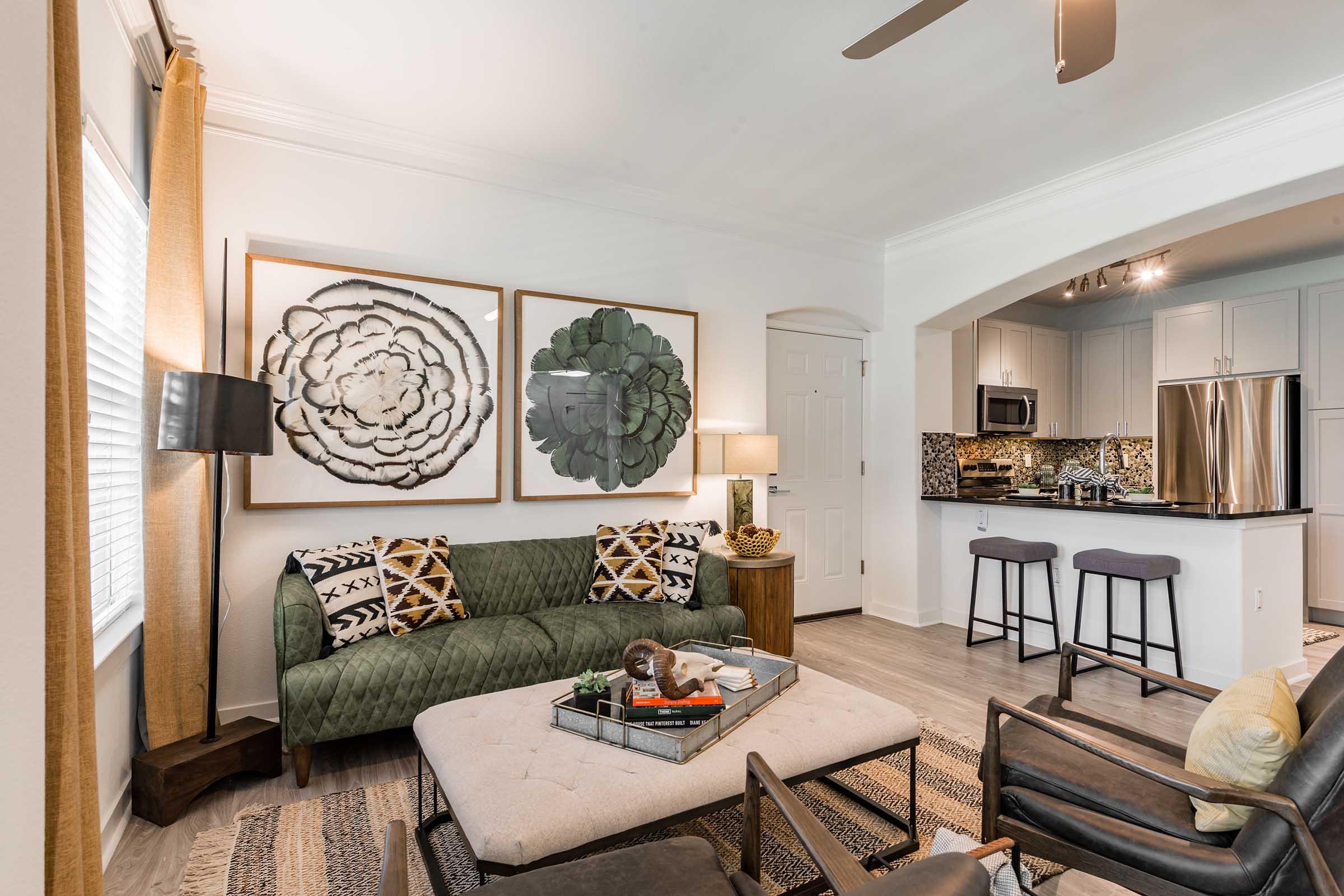 A cozy living room featuring a green velvet sofa with decorative pillows, an ottoman in the center, and two large floral wall art pieces. In the background, there's a modern kitchen with stainless steel appliances and a bar area with two stools. Natural light streams through the window, enhancing the warm atmosphere.