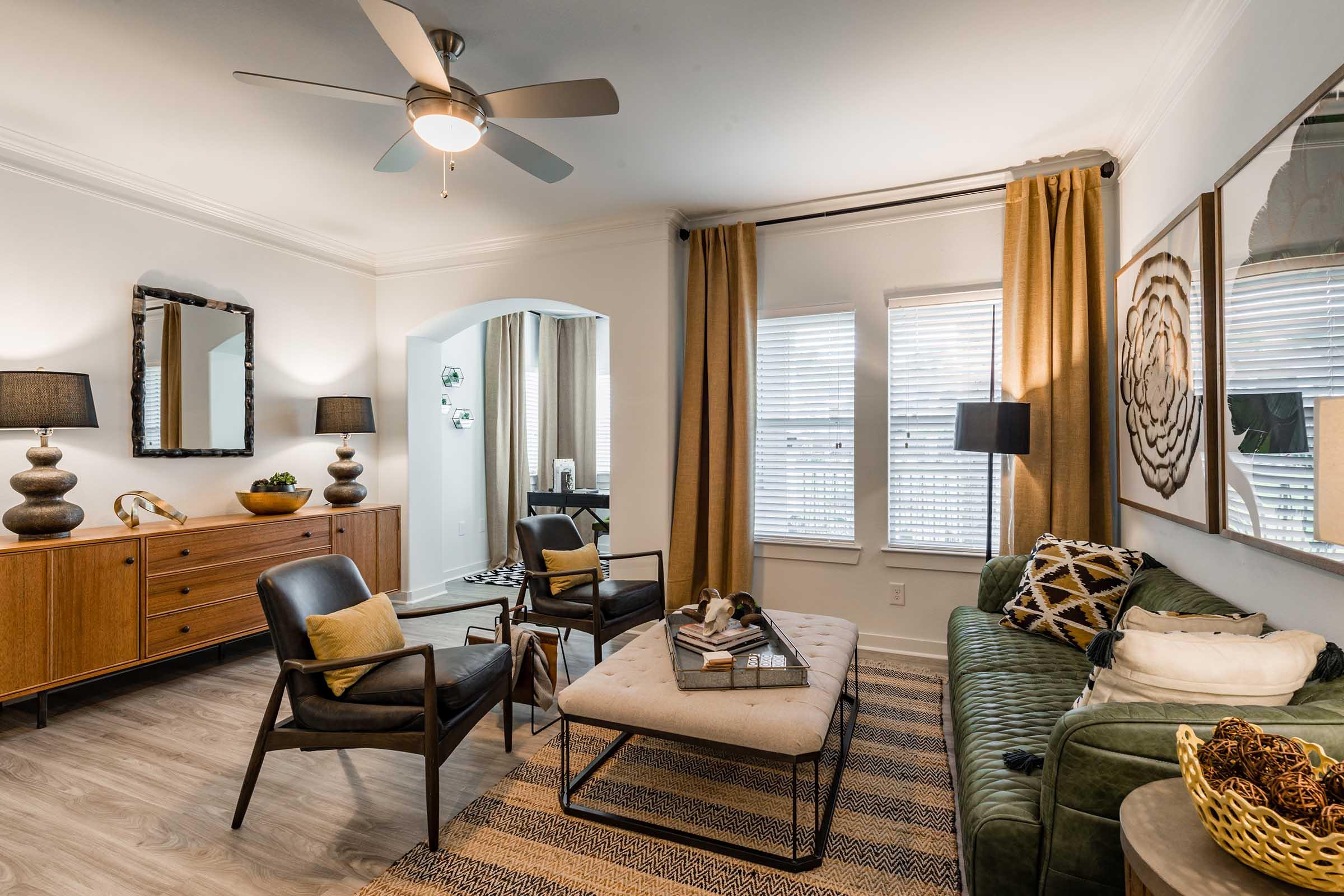 A stylish living room featuring a green sofa with decorative pillows, a coffee table with books and accessories, two black accent chairs, and a warm wooden sideboard. Natural light filters through window treatments. The decor includes modern art, lamps, and a ceiling fan, creating a cozy, inviting atmosphere.