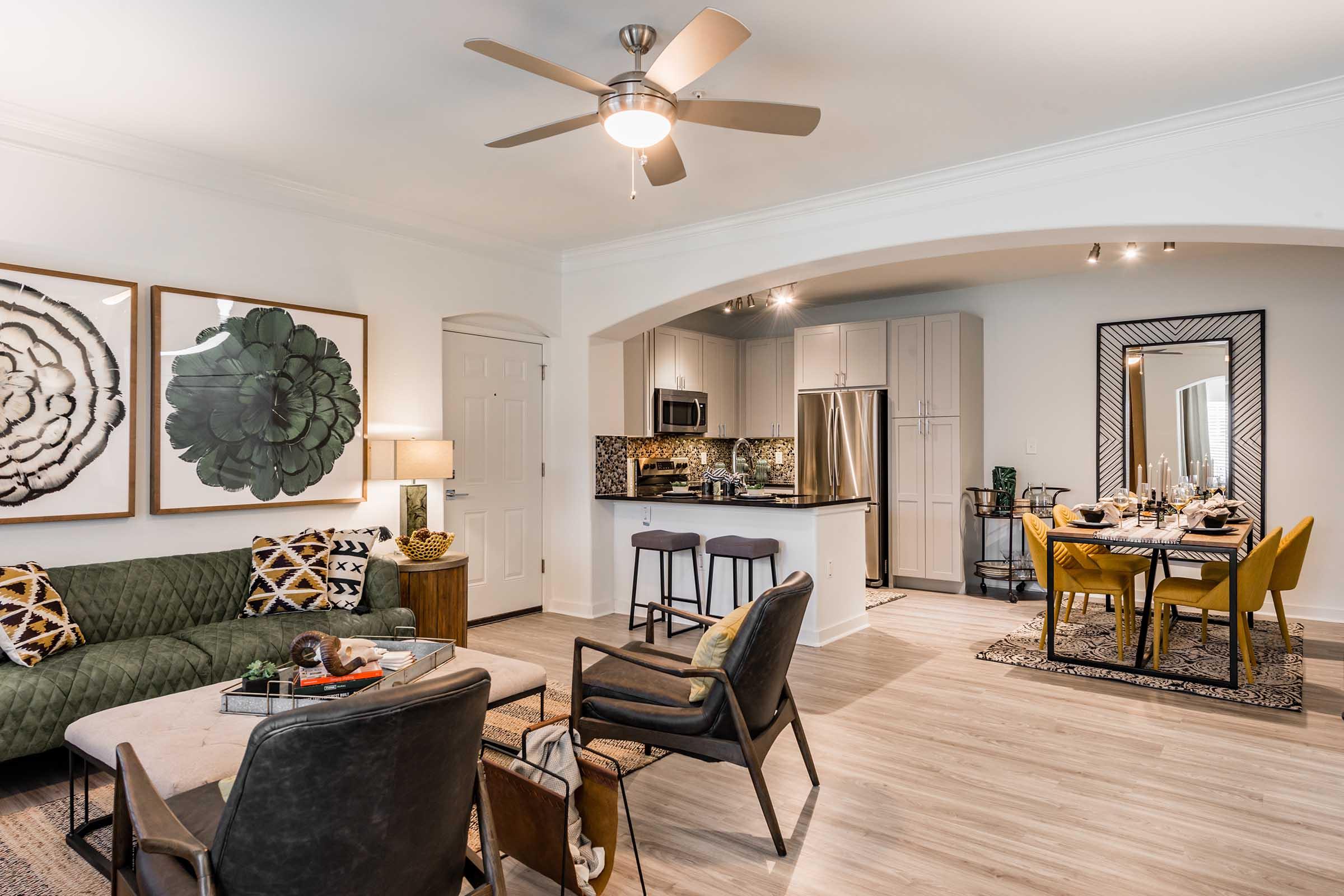 A modern living room featuring a green sectional sofa, stylish armchairs, and a coffee table. The space is decorated with framed botanical art, an overhead ceiling fan, and a dining area with a table set for four. The kitchen area is visible with stainless steel appliances and a decorative backsplash.