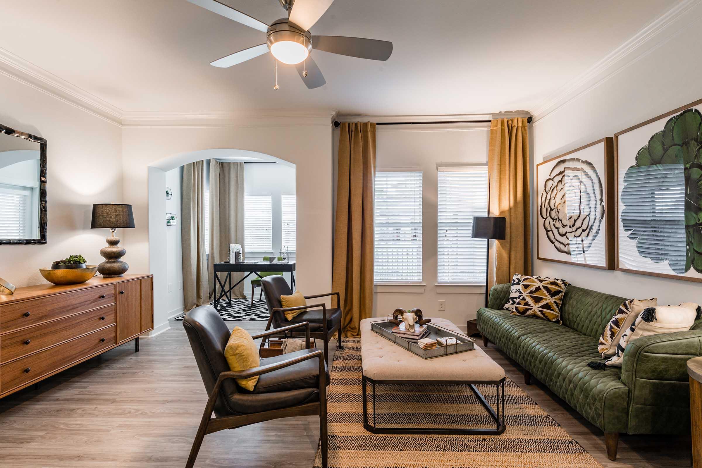 A stylish living room featuring a green velvet sofa, decorative pillows, and a coffee table. There is a wooden sideboard, a large mirror, and two armchairs. The space has a light color palette with mustard curtains and patterned rugs, complemented by natural light from large windows. The decor includes artwork and tasteful accessories.