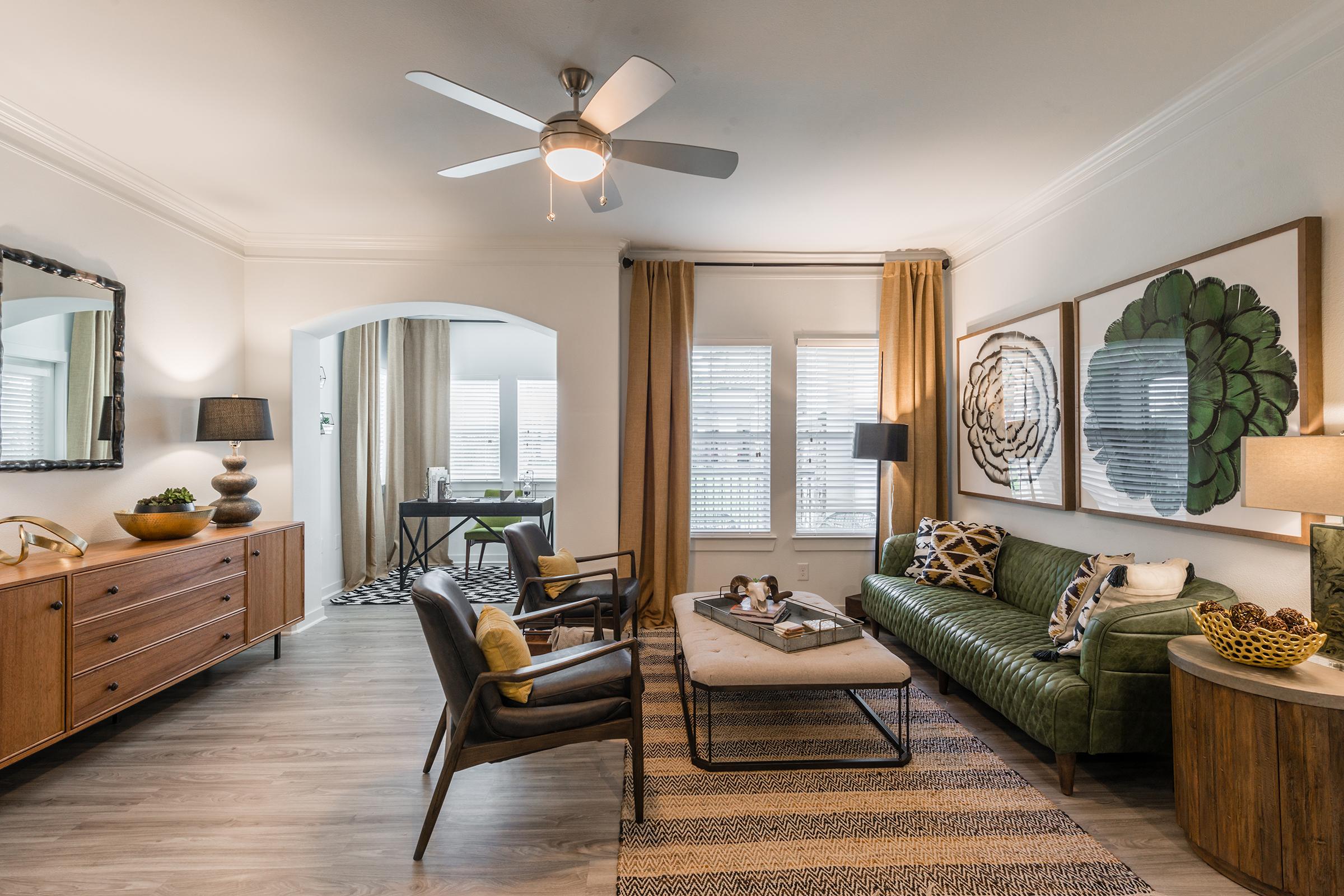 A stylish living room featuring a green velvet sofa, a coffee table with decorative items, and two black armchairs. A wooden sideboard is against the wall, and large windows are adorned with curtains. Decorative wall art adds a modern touch, while a ceiling fan circulates the air in this inviting space.