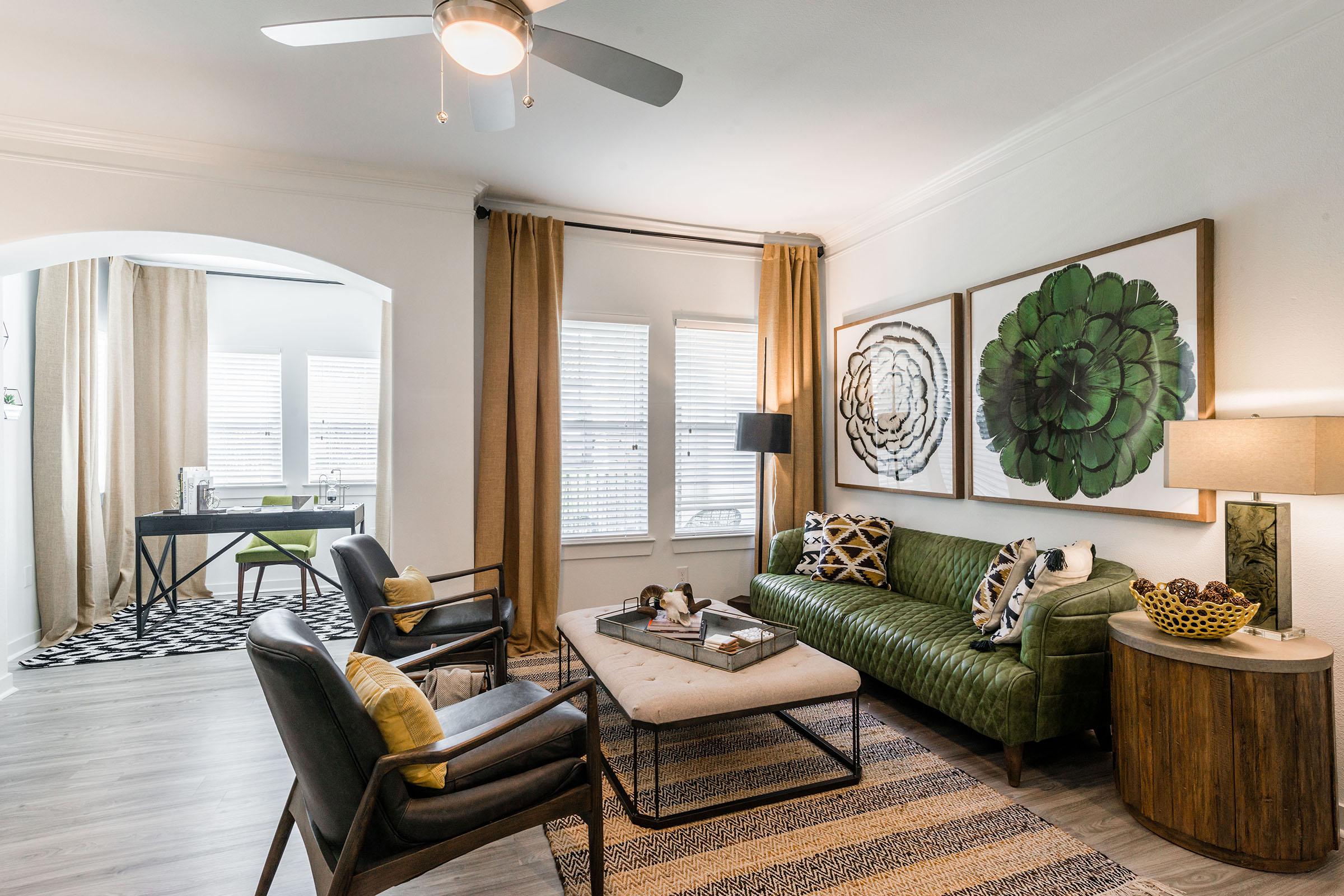 A modern living room featuring a green velvet sofa with decorative pillows, a black and white coffee table, and two armchairs. Large windows with curtains allow natural light. Artwork of green floral designs adorns the walls, and a stylish lamp and decorative elements complete the cozy ambiance.