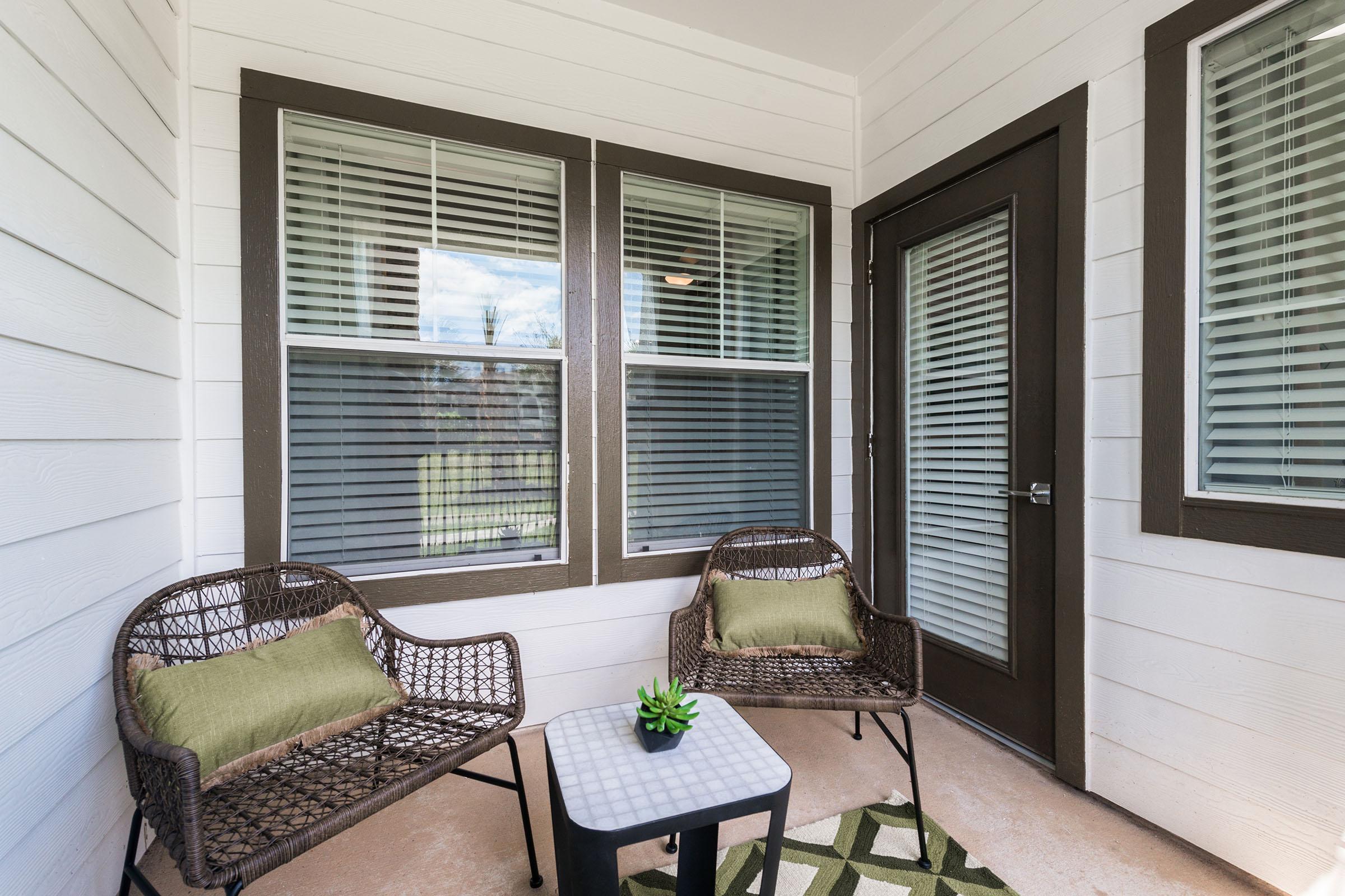 A cozy porch featuring two woven chairs with green cushions and a small square table. The space has white walls with large windows adorned with blinds, allowing natural light to enter. A small potted plant sits on the table, creating a welcoming and peaceful atmosphere.