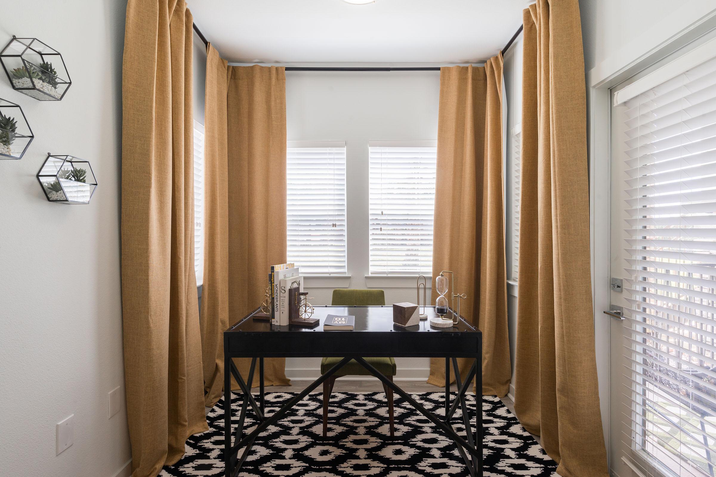 A small, modern home office featuring a black desk with a chair, decorative items, and a small plant. Light-filtering window treatments frame two windows with blinds, and a patterned rug covers the floor. Geometric wall shelves display small succulents, adding a touch of greenery to the space.