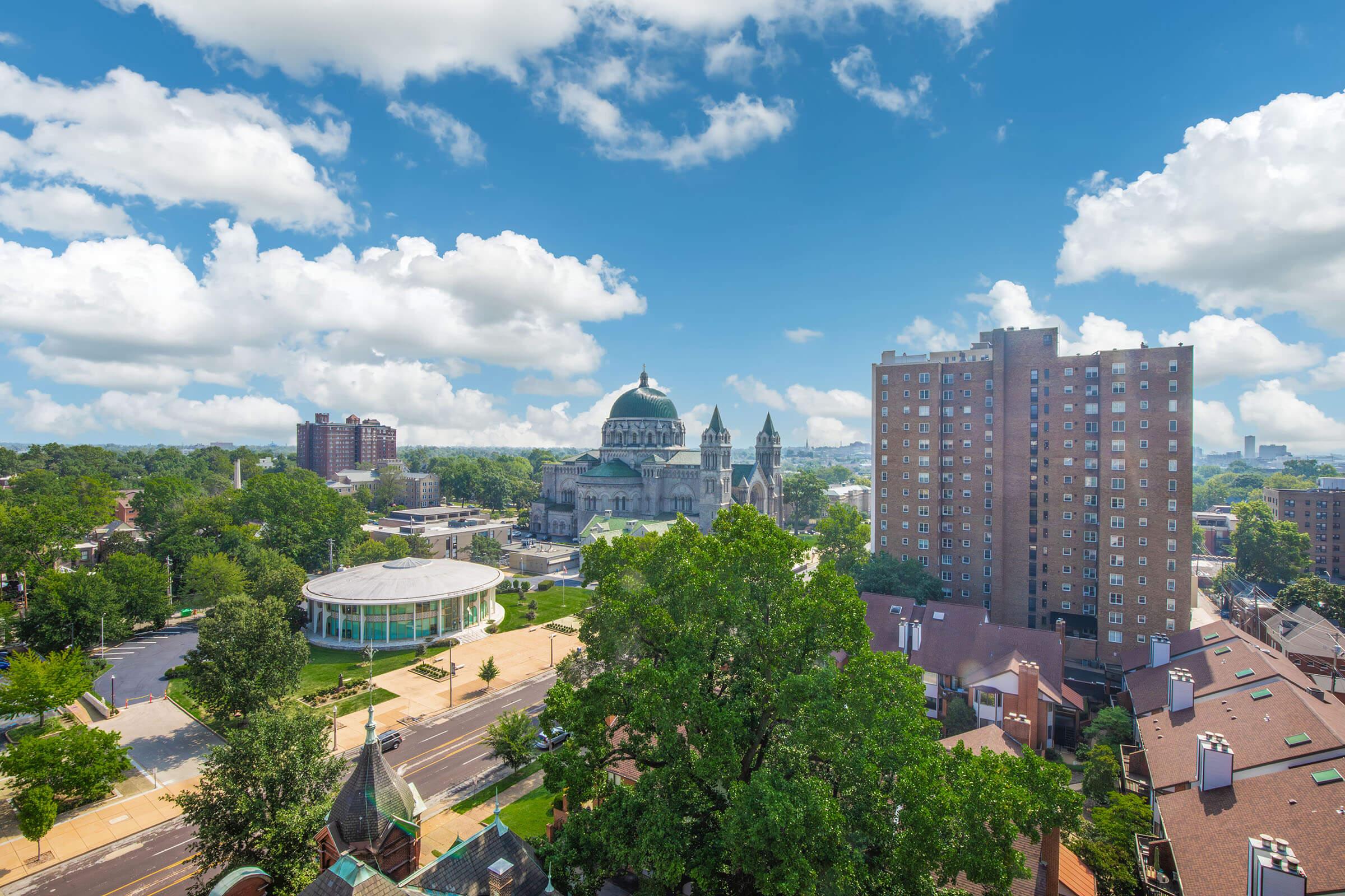 A panoramic view of a cityscape featuring a prominent dome-shaped building surrounded by greenery and modern buildings. In the foreground, tree-lined streets and a circular structure are visible, with a bright blue sky filled with fluffy white clouds overhead.