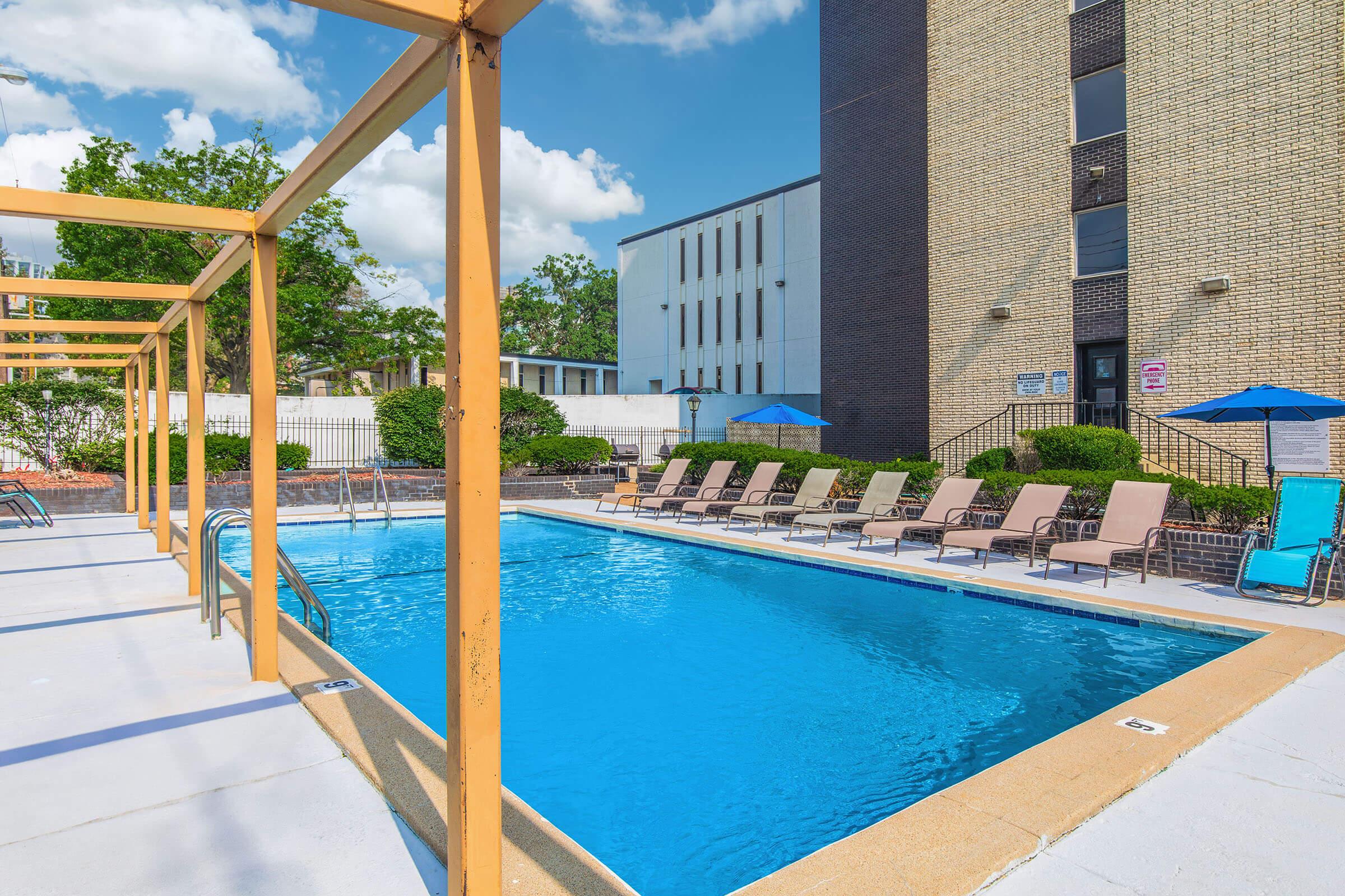 A clear blue swimming pool surrounded by lounge chairs and umbrellas, with a building in the background. The pool area is well-lit by sunlight and features a modern design with potted plants and a clean, inviting atmosphere.