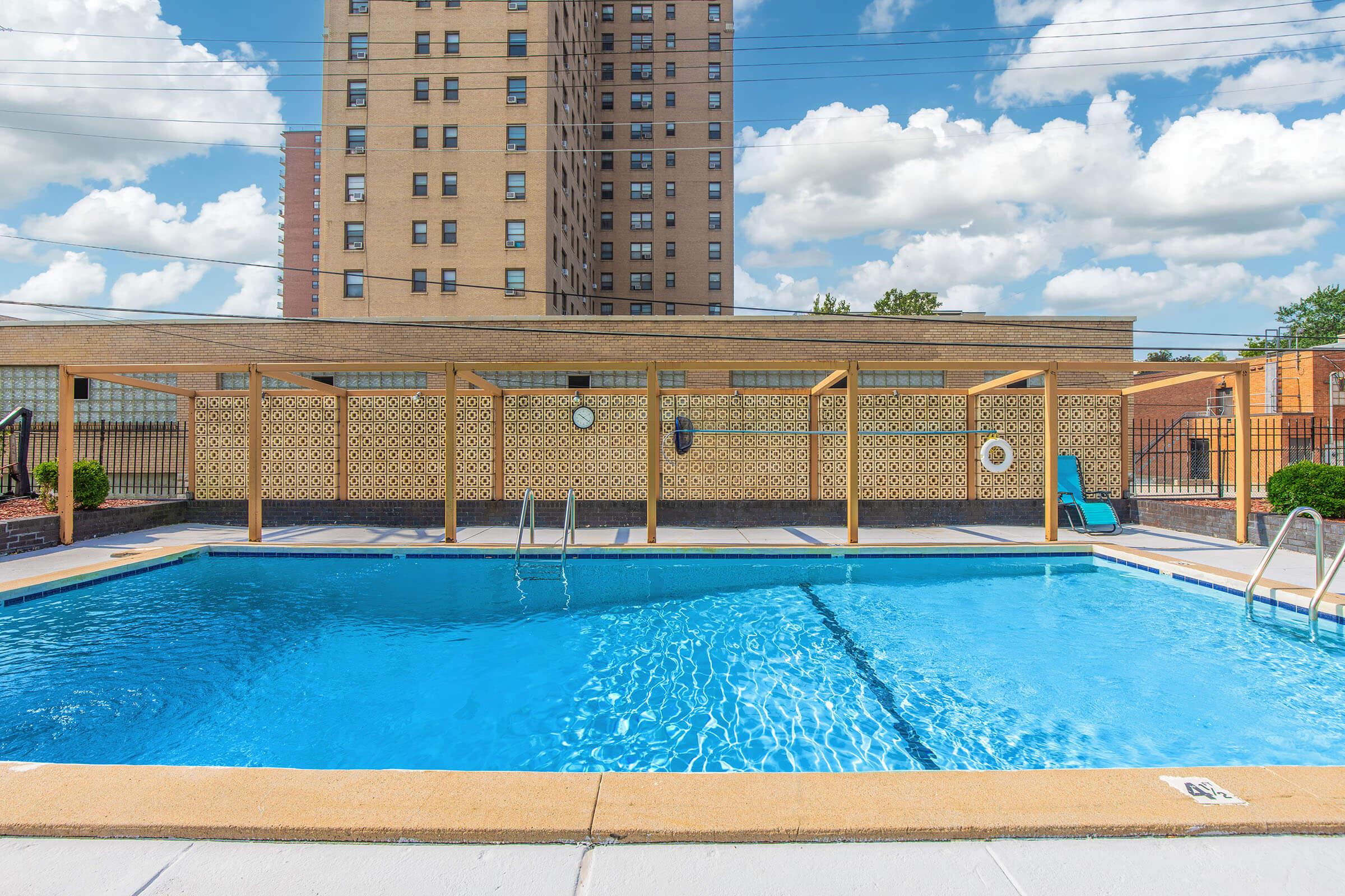 A clear swimming pool surrounded by a wooden pergola, with a few lounge chairs nearby. In the background, a tall residential building and a partly cloudy sky can be seen. The pool area is clean and inviting, making it suitable for relaxation and leisure activities.
