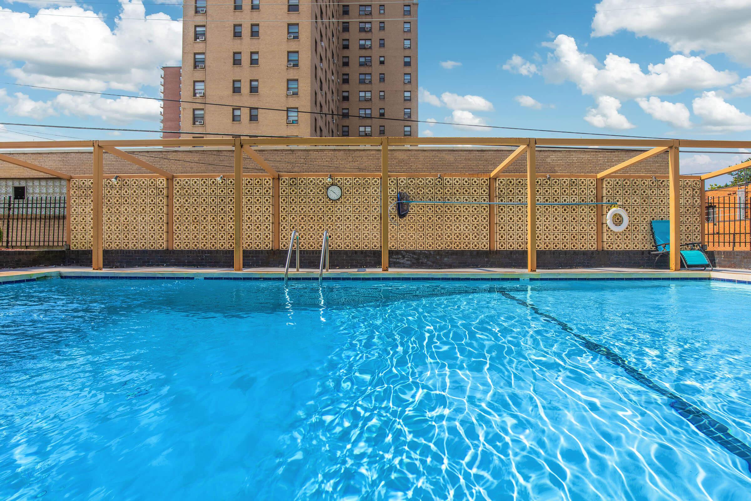 A clear blue swimming pool surrounded by a wooden enclosure, with a tall brick building in the background. The sky is bright with fluffy white clouds. The pool features a ladder for entry, and there are lounge chairs nearby, creating a relaxing atmosphere.