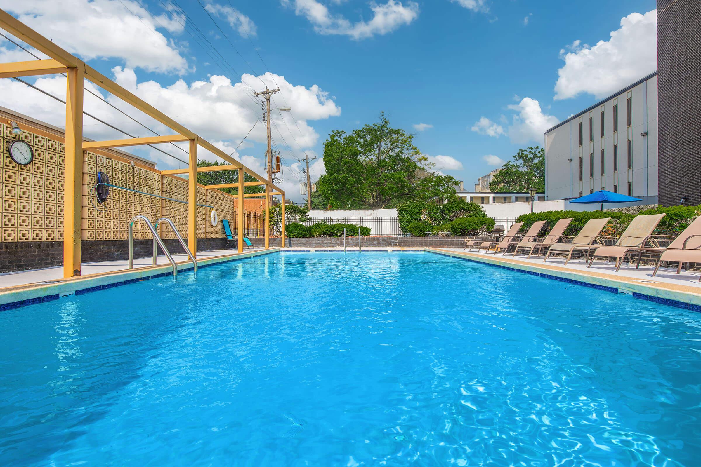 A bright outdoor swimming pool with clear blue water, surrounded by lounge chairs. Wooden decking and decorative walls add style, while fluffy white clouds dot a vibrant blue sky. Nearby buildings and greenery complete the inviting atmosphere.