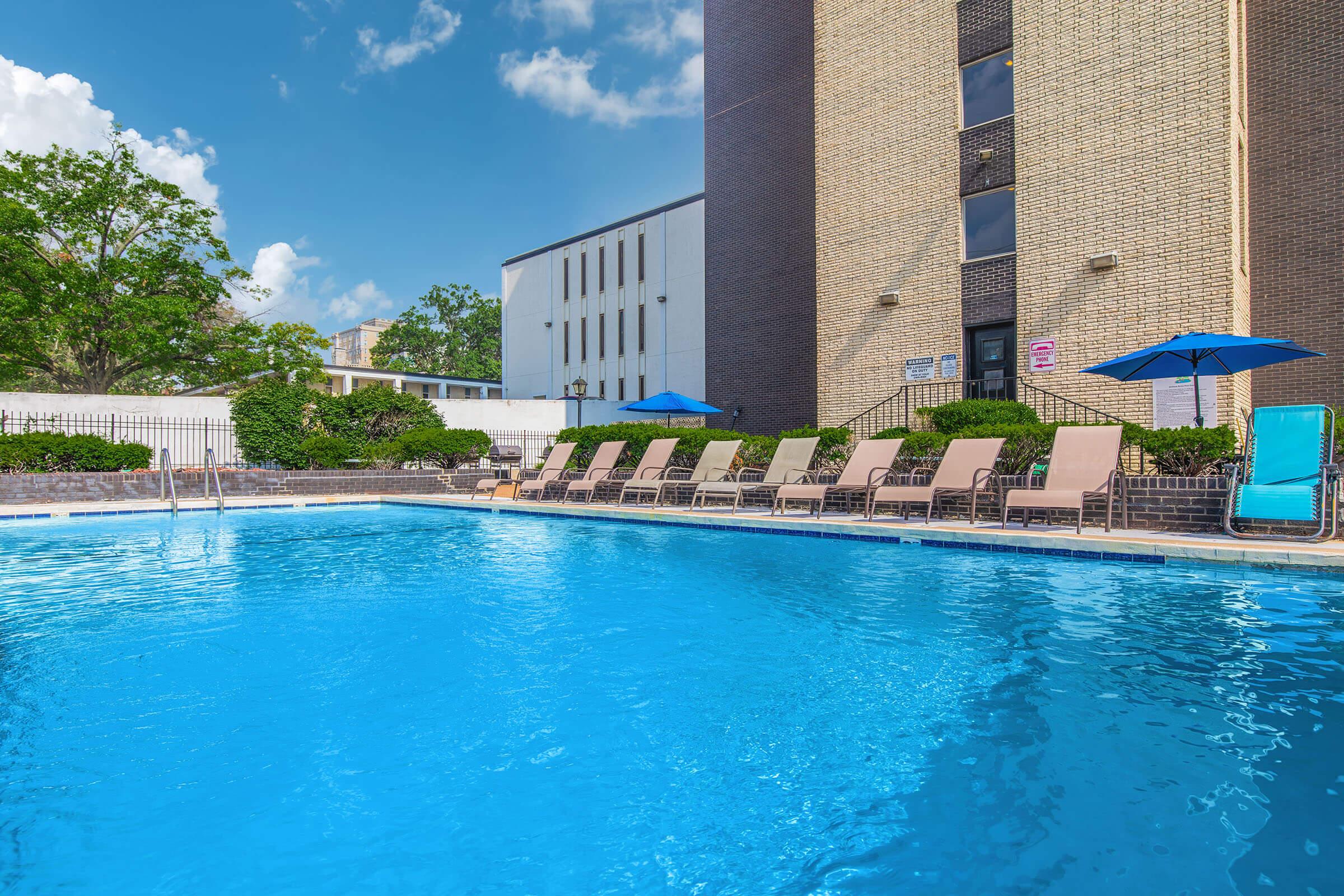 A sparkling blue swimming pool surrounded by lounge chairs and umbrellas, with a brick building in the background. The scene is set against a bright blue sky with a few fluffy clouds, creating a relaxing atmosphere ideal for leisure and enjoyment.