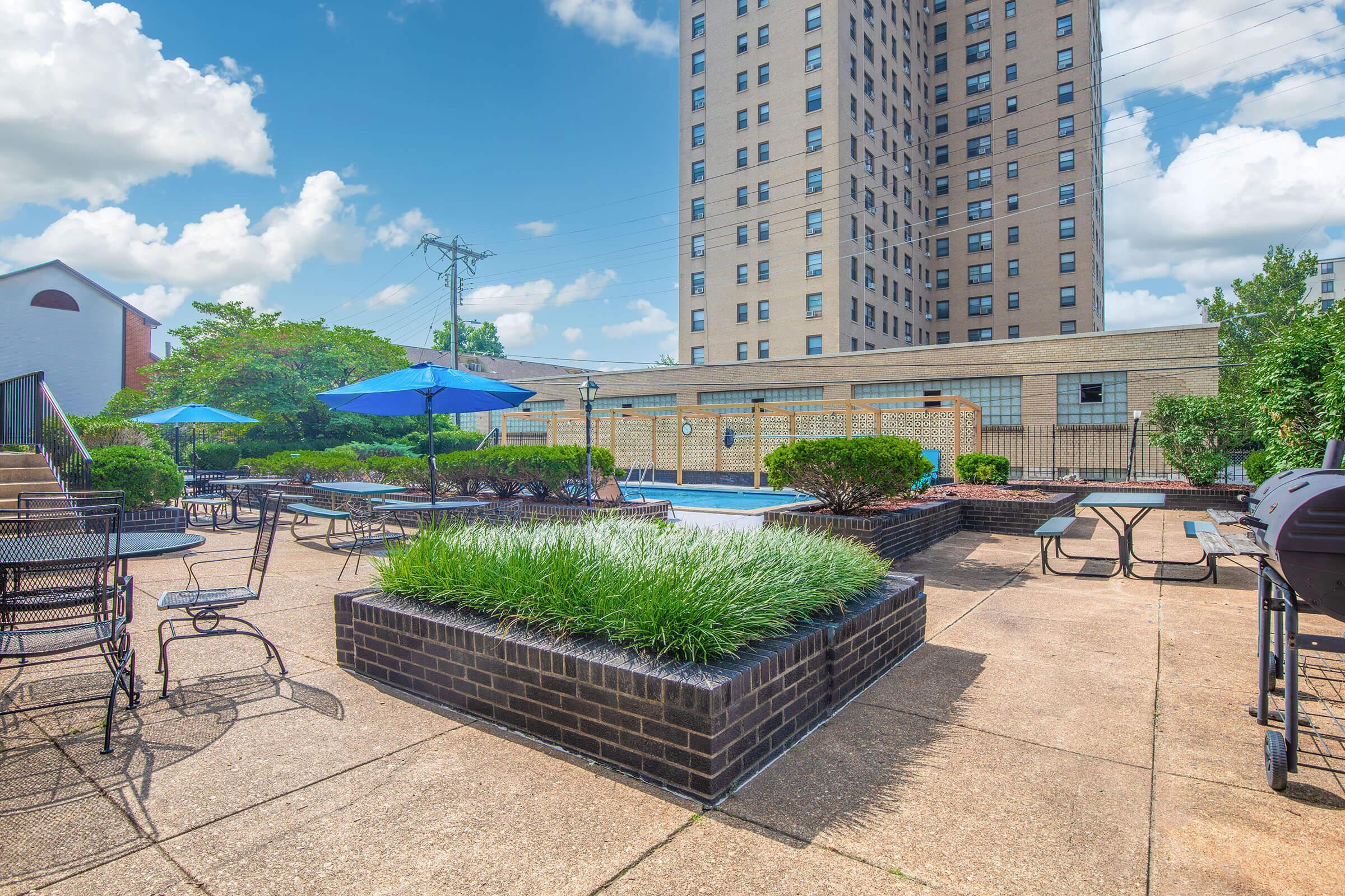 A sunny outdoor area featuring a swimming pool surrounded by green bushes and neatly trimmed grass. Several blue umbrellas are set up over tables and chairs, providing shaded seating. A high-rise building is visible in the background under a partly cloudy sky.