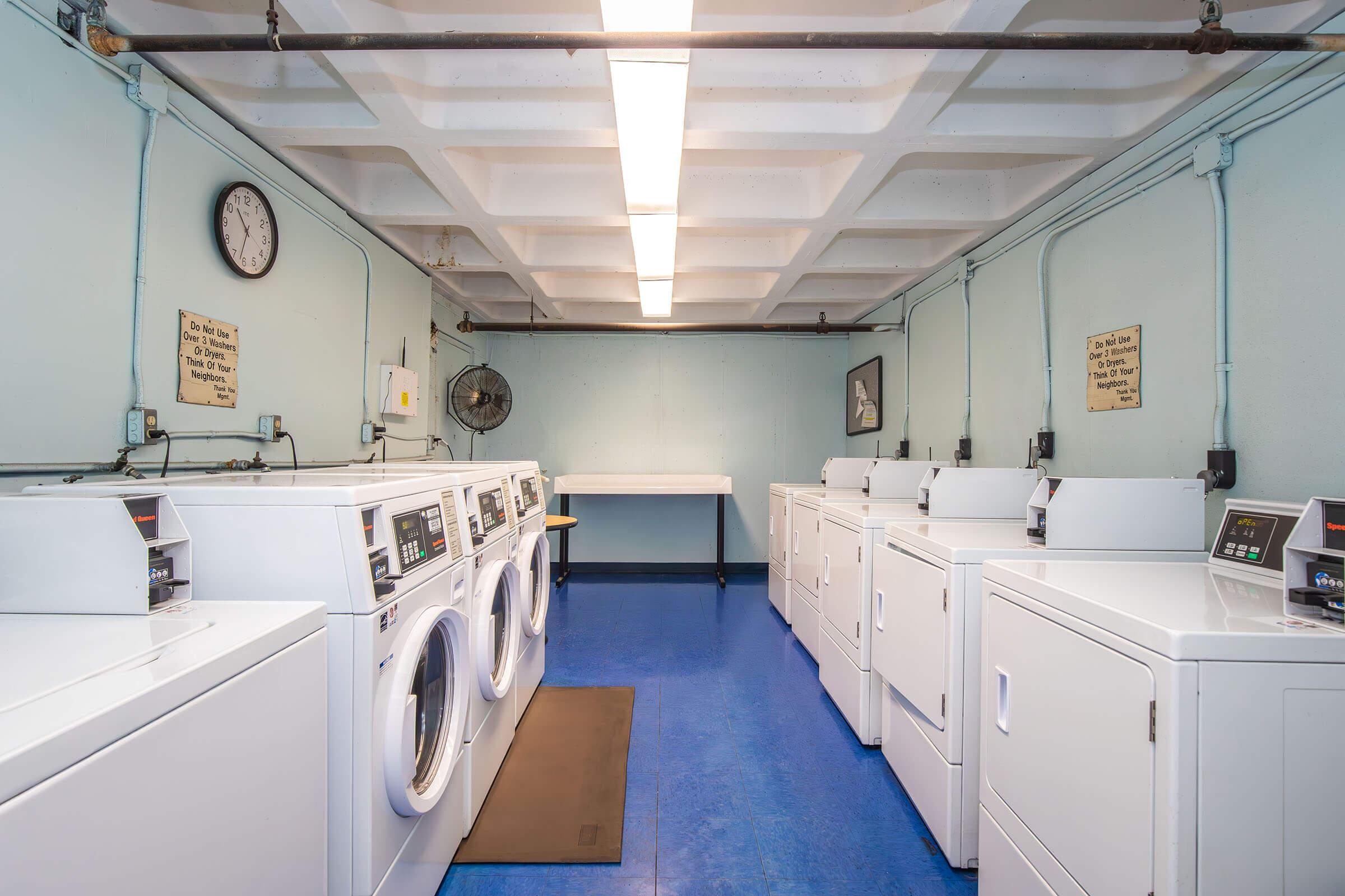 A clean and well-lit laundromat featuring multiple white washing machines and dryers lined up against light blue walls. A clock is mounted on the wall, and there are two framed pictures. The floor is blue, and a folding table is positioned at the far end of the room.
