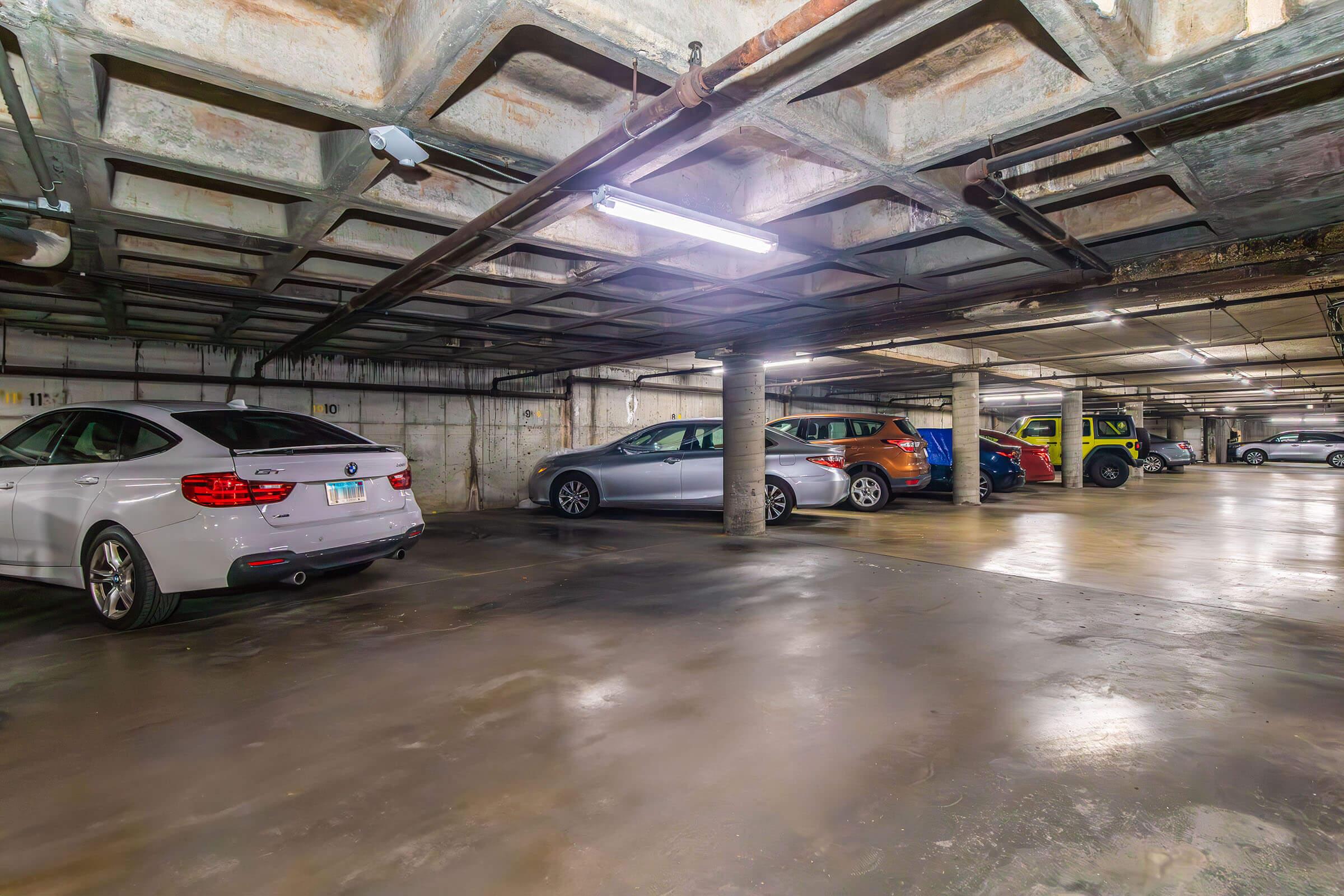 A dimly lit underground parking garage featuring several parked cars, including a silver sedan, an orange SUV, a gray sedan, and a bright green vehicle. The concrete walls and ceiling display industrial textures, and overhead lighting casts a soft glow over the scene.