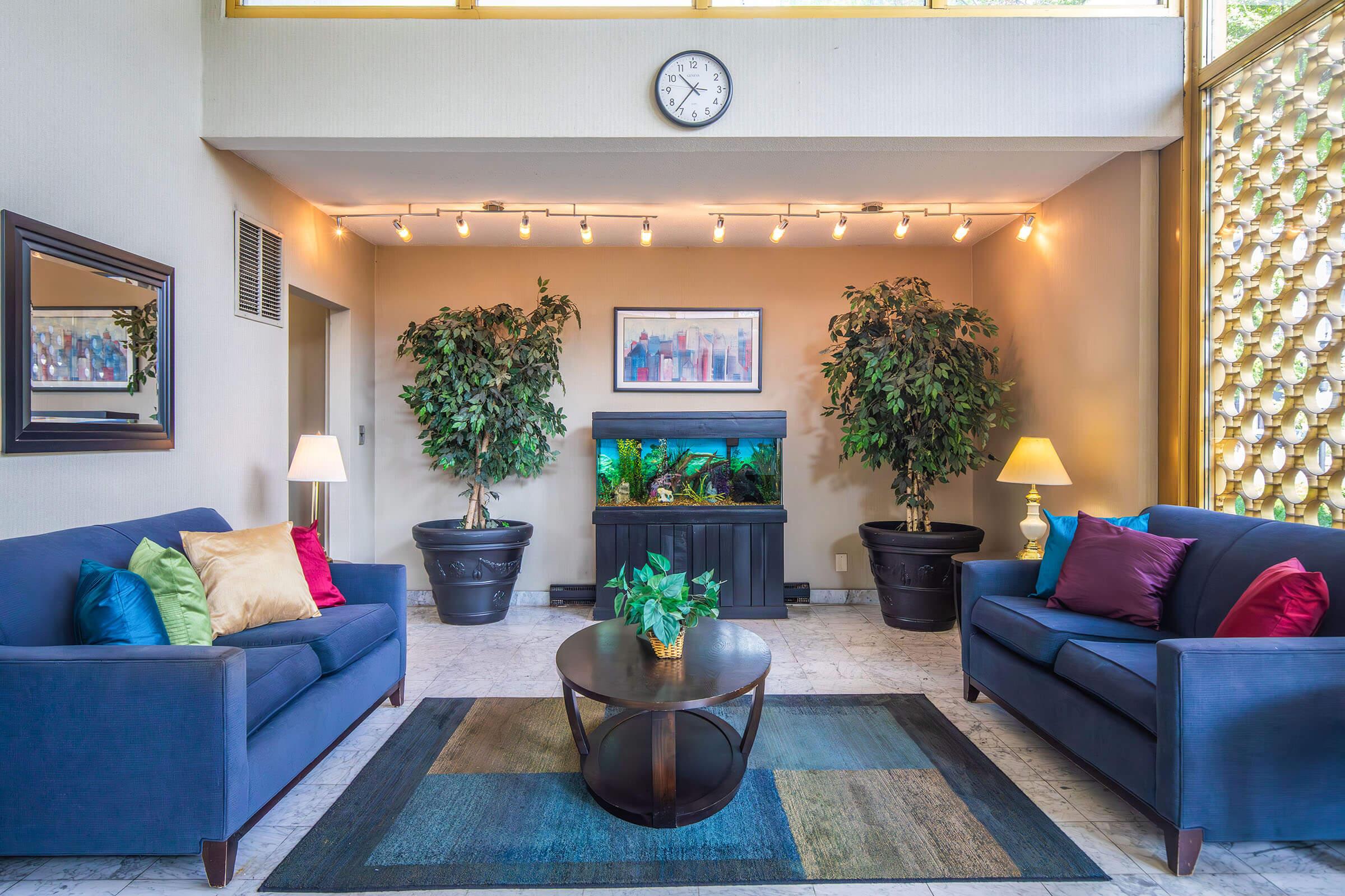 A modern lounge area featuring two blue sofas with colorful cushions, a round coffee table, and a planter with greenery. A fish tank serves as a focal point, surrounded by potted plants. Soft lighting is provided by wall-mounted fixtures, and a clock hangs above. The space has a clean, contemporary design.