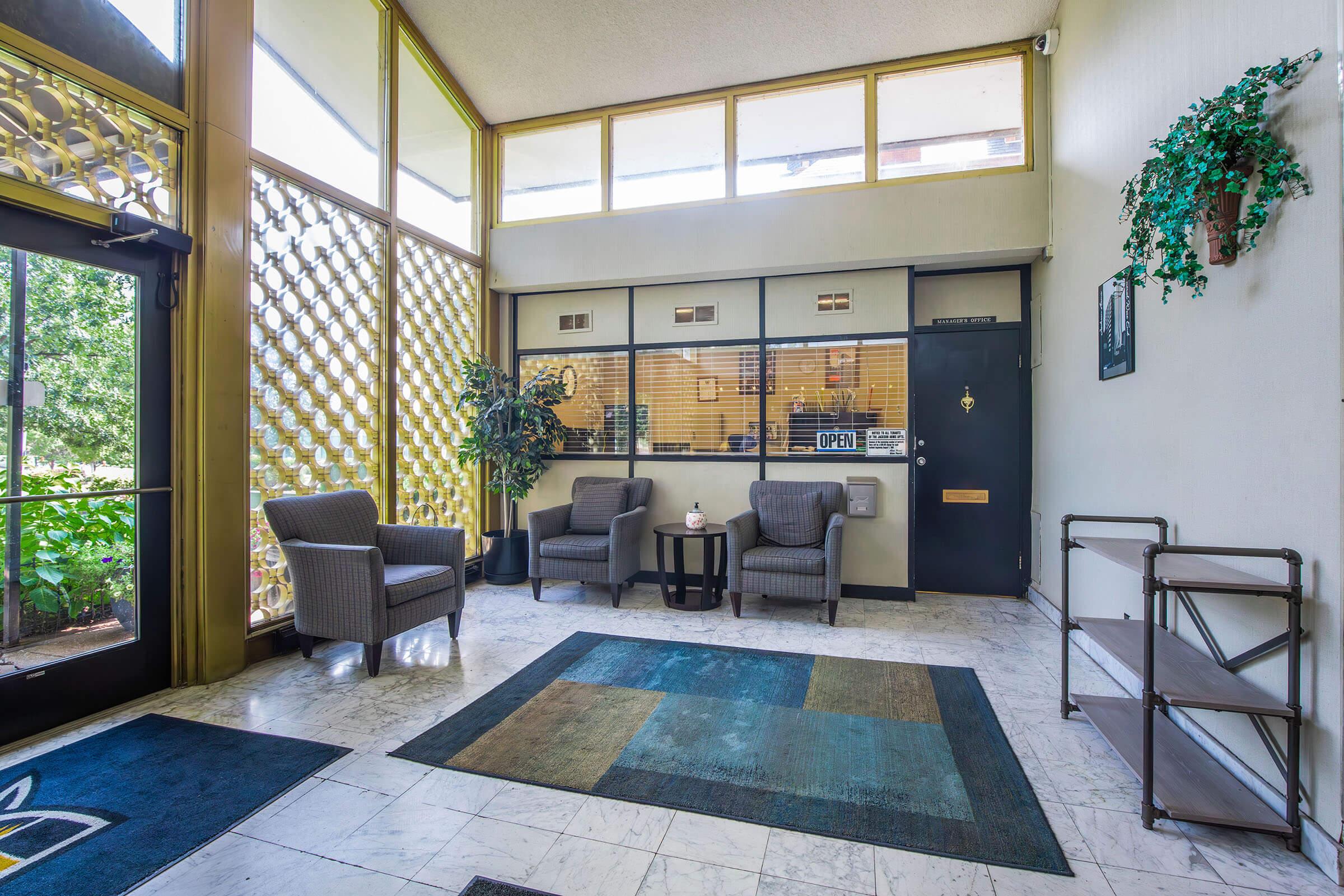 A lobby area featuring comfortable gray chairs, a small table with a flower vase, and a decorative plant. There's a large window allowing natural light in, and a patterned brown and blue rug on the marble floor. A reception desk with an "OPEN" sign is visible, along with a wall-mounted shelf.
