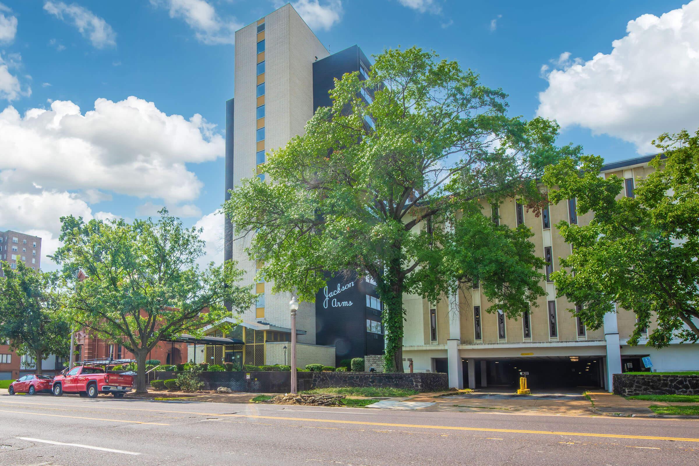 A view of a multi-story hotel or apartment building with a modern design, surrounded by large trees and greenery. In the foreground, a red car is parked along the street, while a parking garage entrance is visible. The sky is blue with scattered clouds.