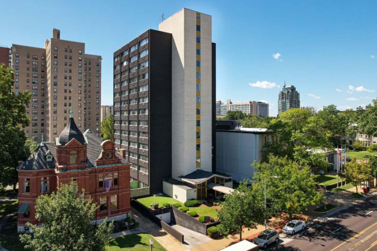 A view of a cityscape featuring a mix of architectural styles, including a historic multi-story red brick building with decorative features in the foreground and a modern high-rise building in the background. Green trees and landscaped areas enhance the urban setting under a clear blue sky.
