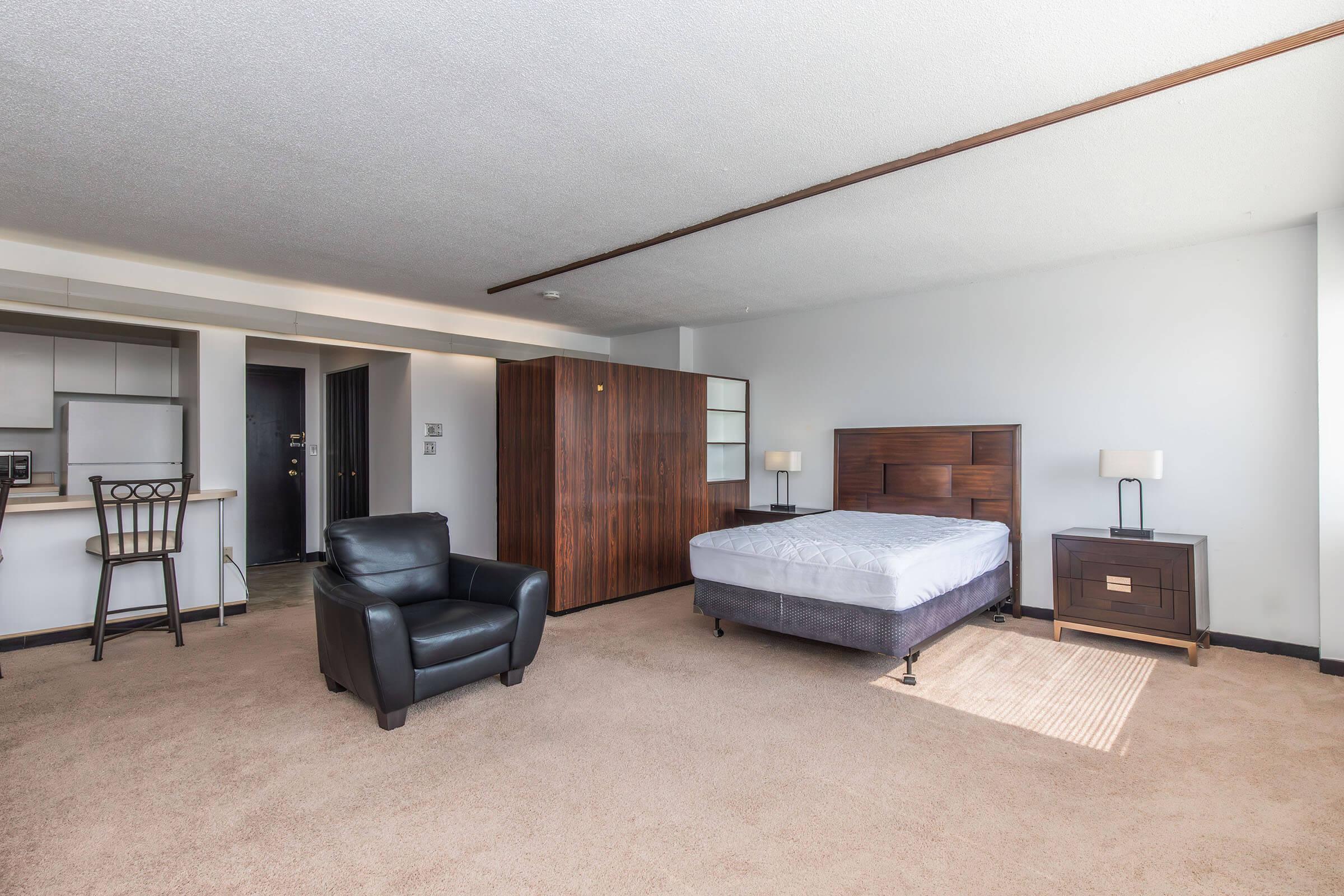 A spacious and well-lit bedroom featuring a queen-sized bed with a dark wood frame, a nightstand with a lamp, a black leather armchair, and a kitchenette area visible in the background. The carpeted floor and neutral-colored walls create a warm and inviting atmosphere.