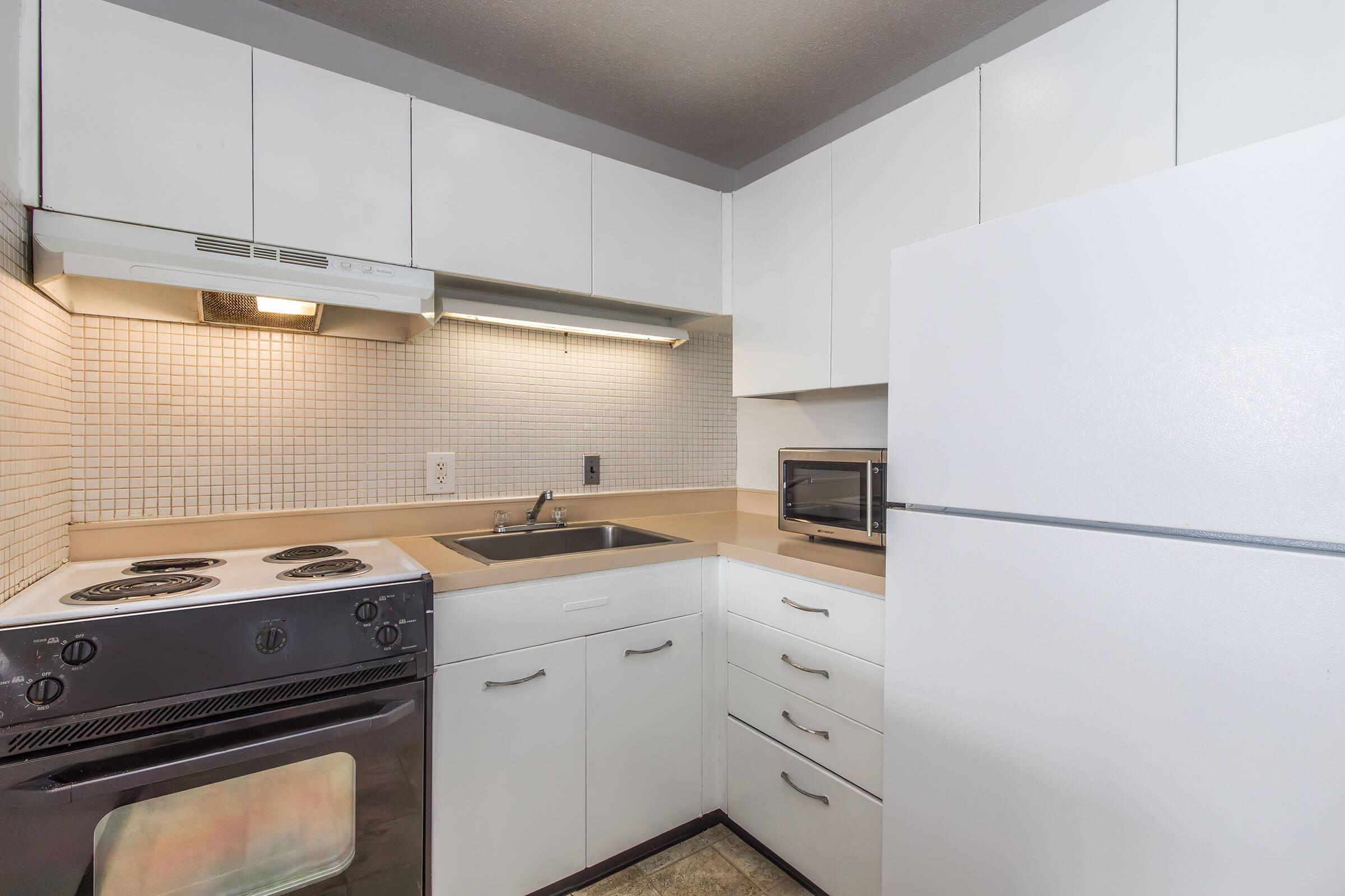 A modern kitchen featuring white cabinets, a black stovetop with an oven below, a sink, a countertop, a microwave, and a white refrigerator. The walls are adorned with light-colored tiles, and the lighting is bright, creating a clean and inviting atmosphere.