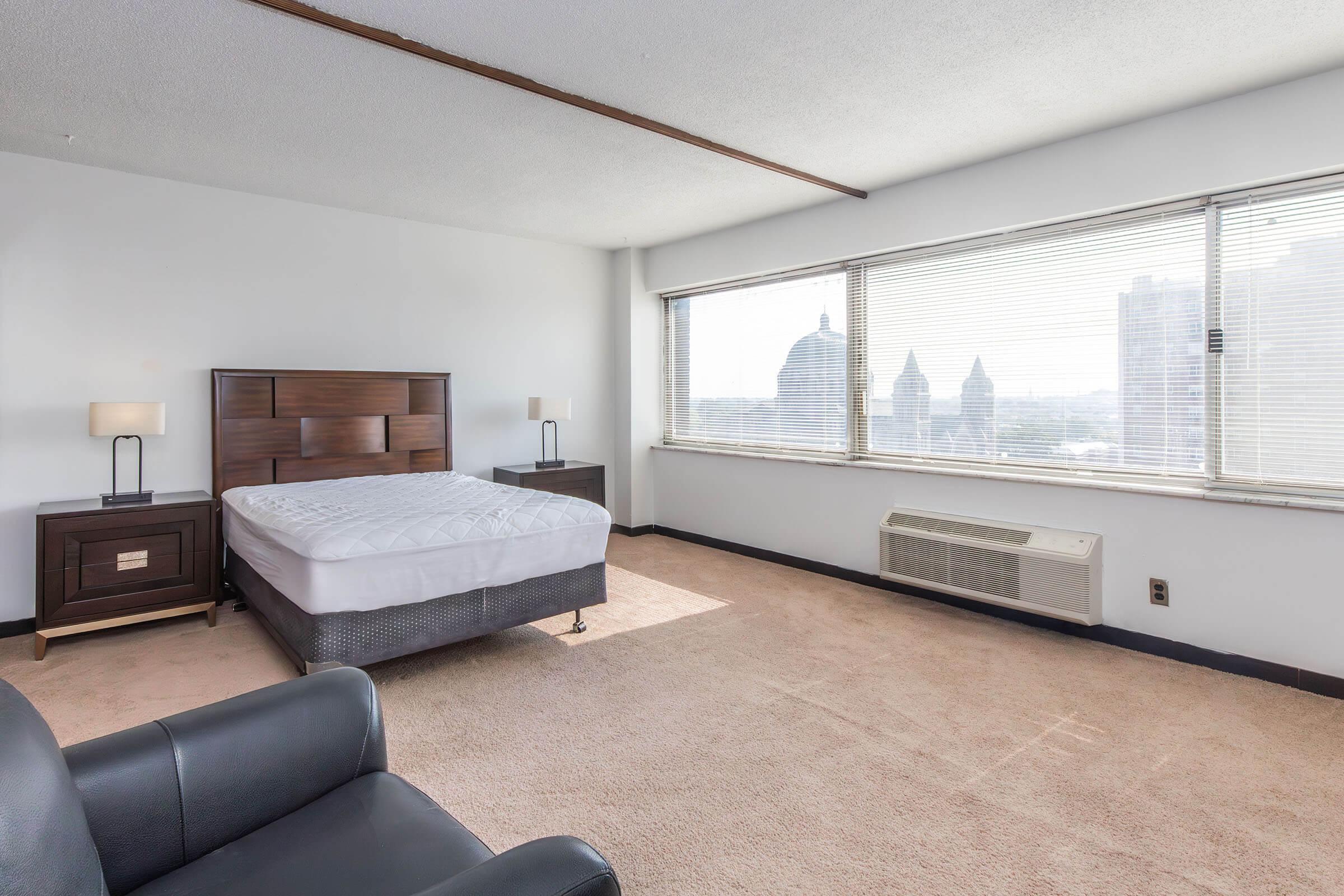 A spacious hotel room featuring a large window with city views, a comfortable bed with a white mattress, a dark wood headboard, and two bedside tables with lamps. A black leather armchair is placed to the side, and the room has light-colored carpet and is well-lit by natural light.
