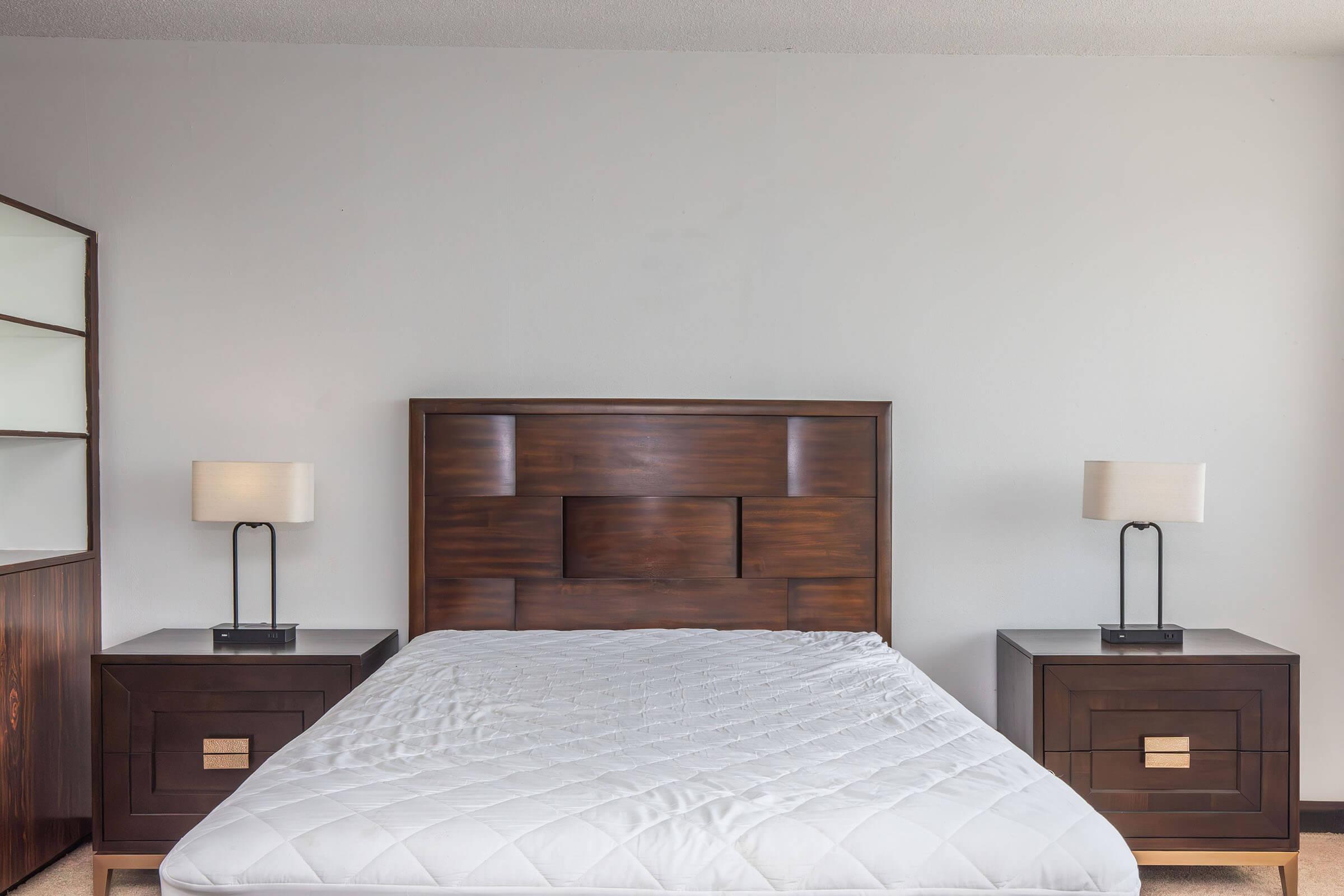 A simple bedroom featuring a wooden bed frame with a headboard, two matching bedside tables with modern lamps, and a light gray wall behind. The bed is unmade with no bedding, creating a minimalist aesthetic.