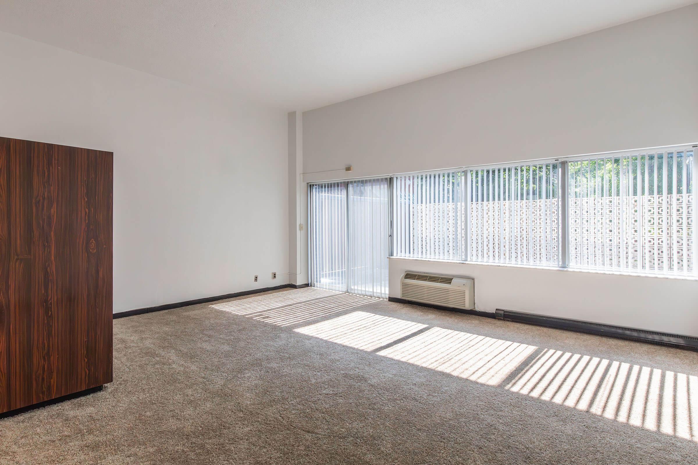 A spacious, empty room with beige carpet and large vertical blinds letting in sunlight. There is a brown wooden cabinet in the corner and an air conditioning unit on the wall. The walls are painted white, and the windows show a view of an exterior wall with patterned design.