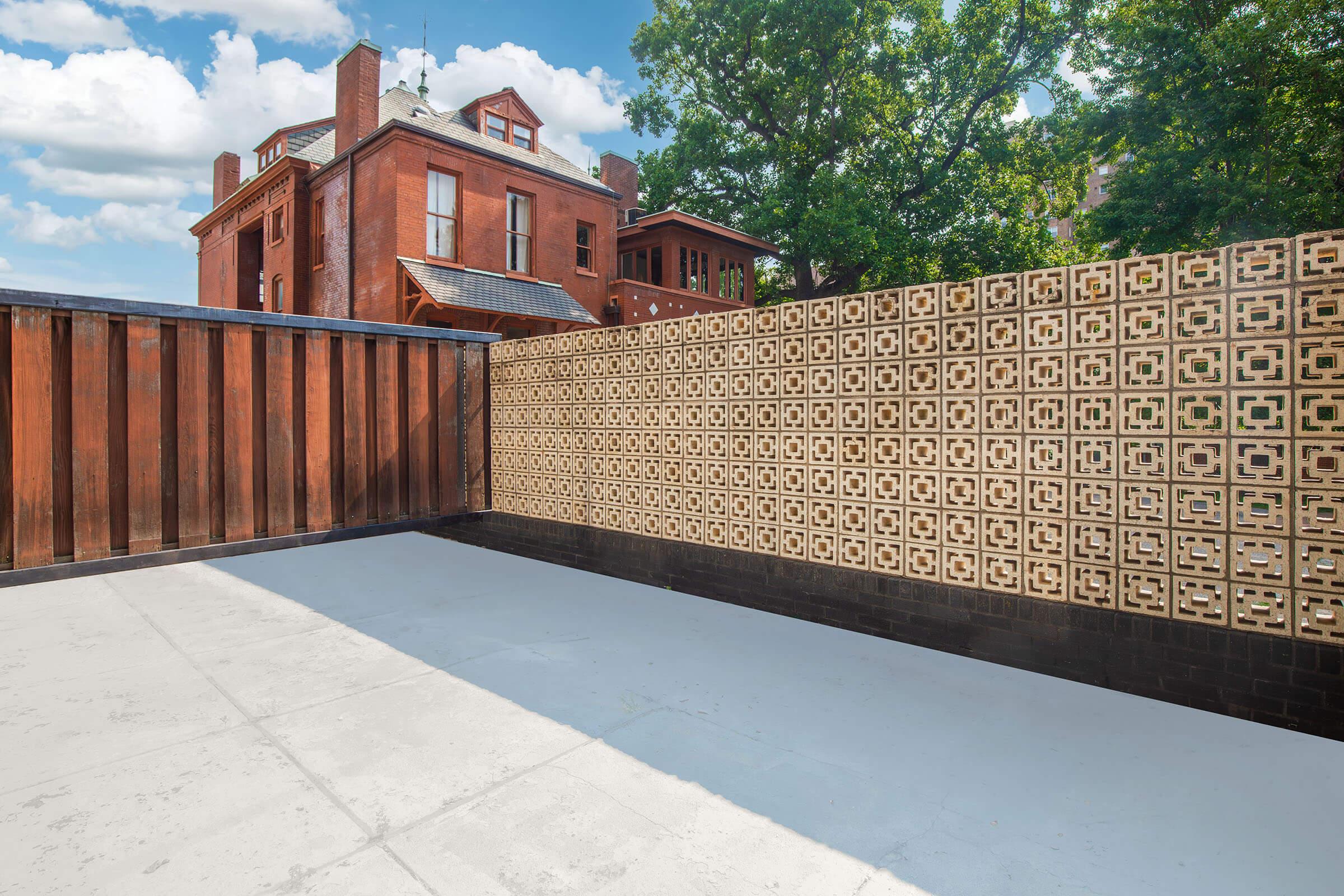 A rooftop terrace featuring a modern design with a patterned brick wall and a wooden barrier. In the background, a historic red brick building is visible under a partly cloudy sky, surrounded by trees. The floor is plain and light-colored, emphasizing the architectural elements of the space.