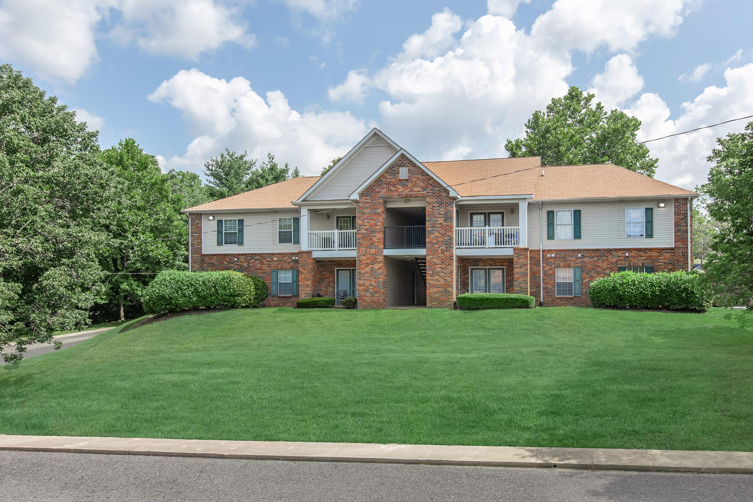 A two-story brick apartment building with white siding is surrounded by neatly trimmed green lawns and bushes. The building features a central staircase leading to a second-floor balcony. The sky is partly cloudy, adding a bright atmosphere to the scene.