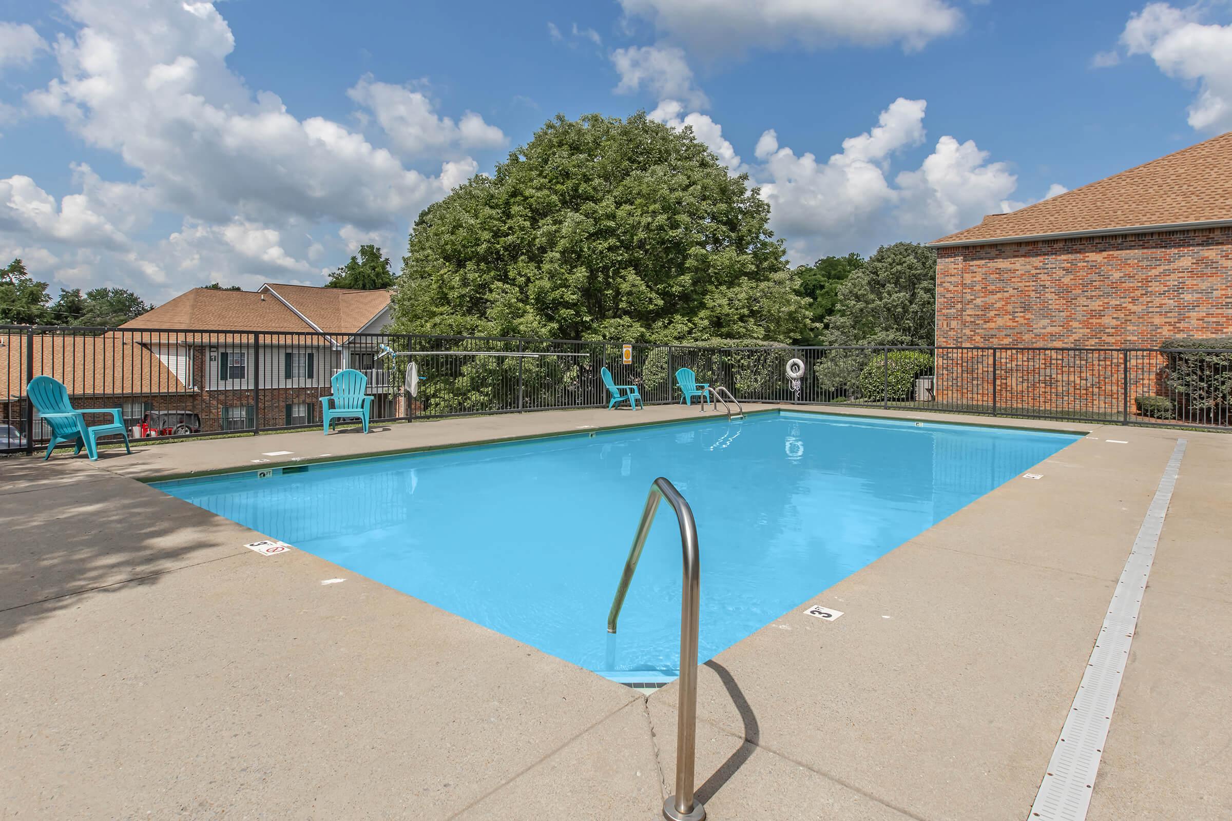 A clear swimming pool surrounded by a deck featuring turquoise lounge chairs, set against a backdrop of green trees and a blue sky with fluffy white clouds. The pool area is part of a residential complex, with brown brick buildings visible nearby.