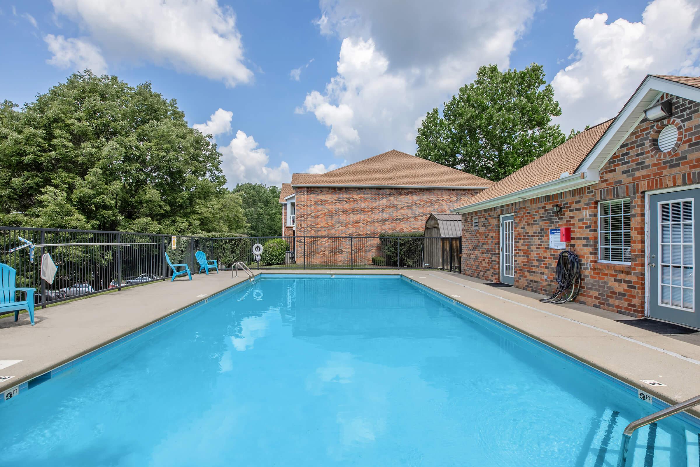A swimming pool surrounded by a concrete deck, with several blue lounge chairs positioned nearby. Lush green trees are visible in the background, along with a brick building. The sky is partly cloudy, creating a bright and inviting atmosphere.