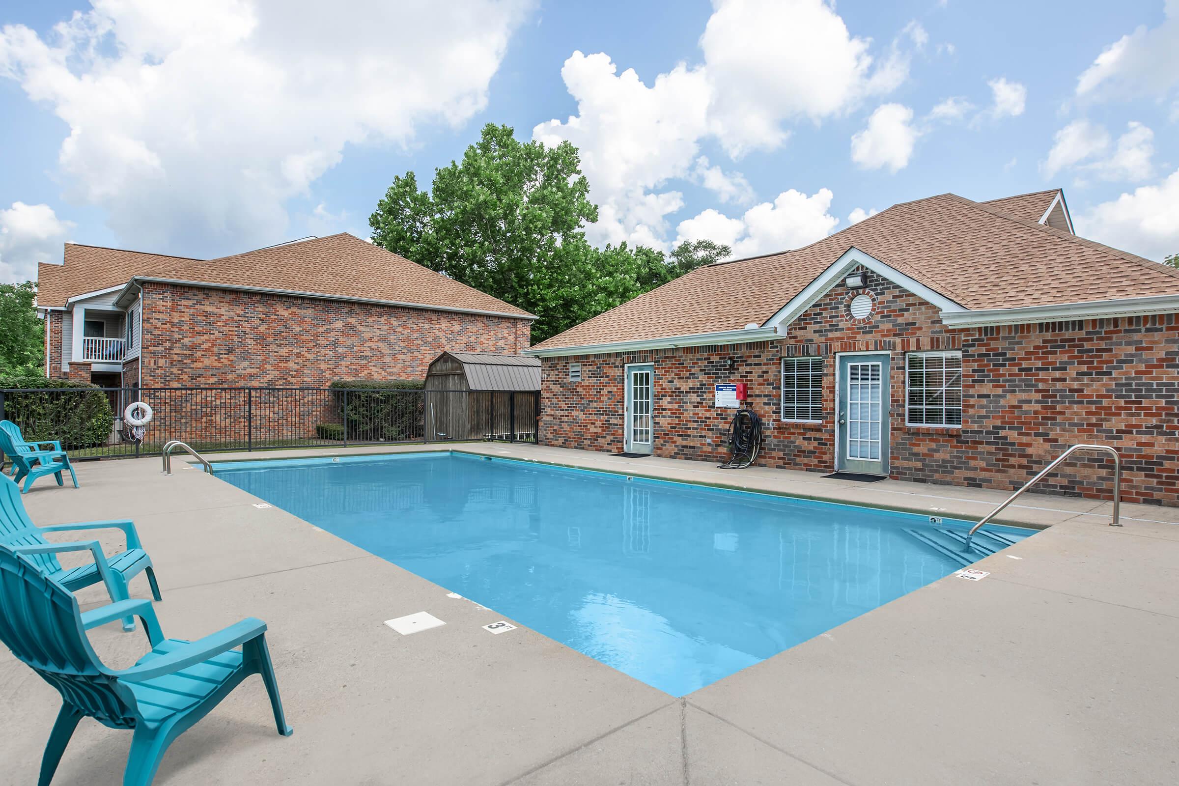 A clear blue swimming pool surrounded by concrete, with green chairs placed around it. A brick building with multiple windows and a sloped roof is visible in the background, along with a shaded area. The sky is partly cloudy, creating a bright and inviting atmosphere.