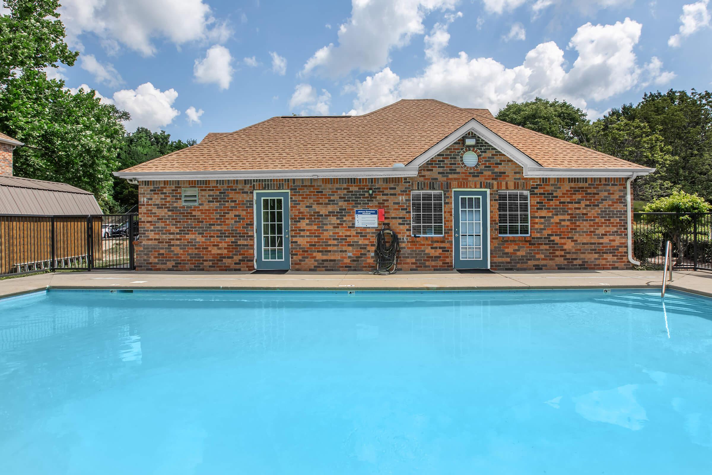 A clear blue swimming pool in front of a brick building with a brown roof. The building features two sets of double doors and a clock on the front. The surrounding area is lush with greenery and the sky is partly cloudy, creating a sunny atmosphere ideal for relaxation and leisure.
