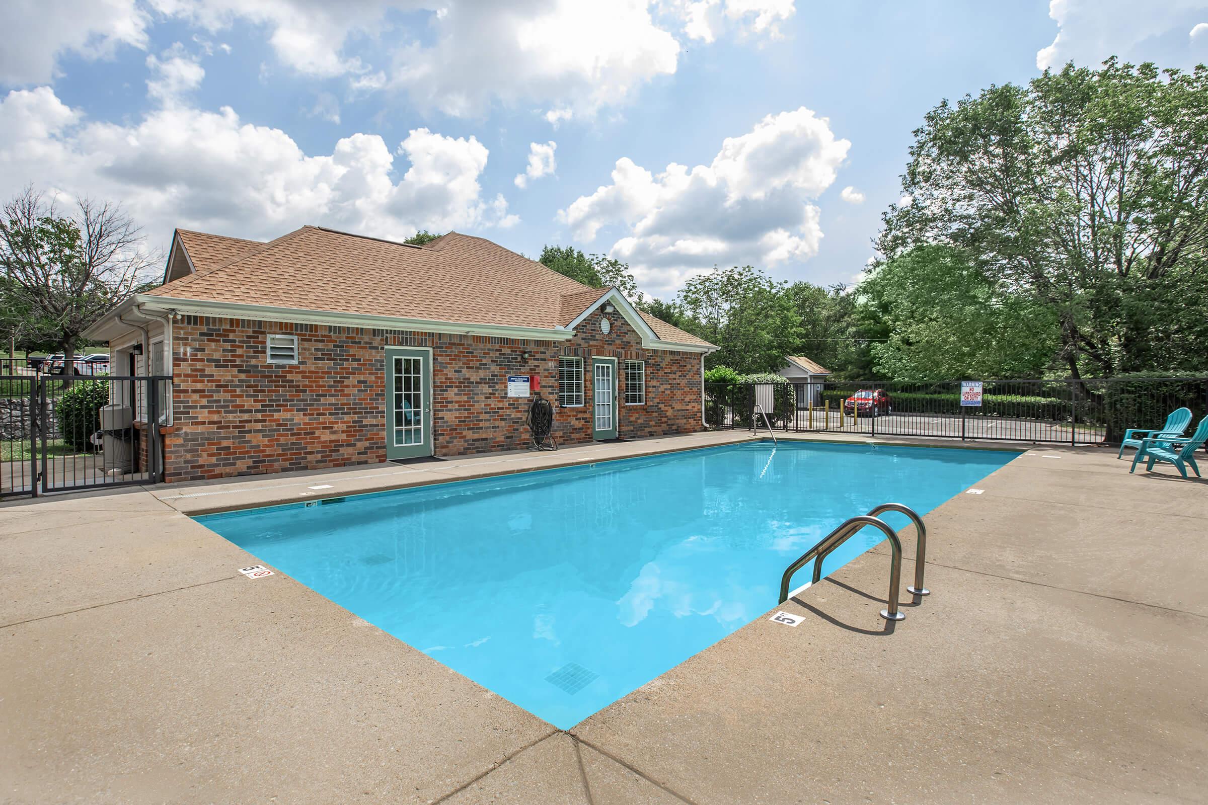 A bright outdoor swimming pool with a clear blue water, surrounded by a concrete deck. In the background, there is a brick building with a sloped roof, featuring large windows and a patio area. Lush green trees and a cloudy sky complete the serene atmosphere.