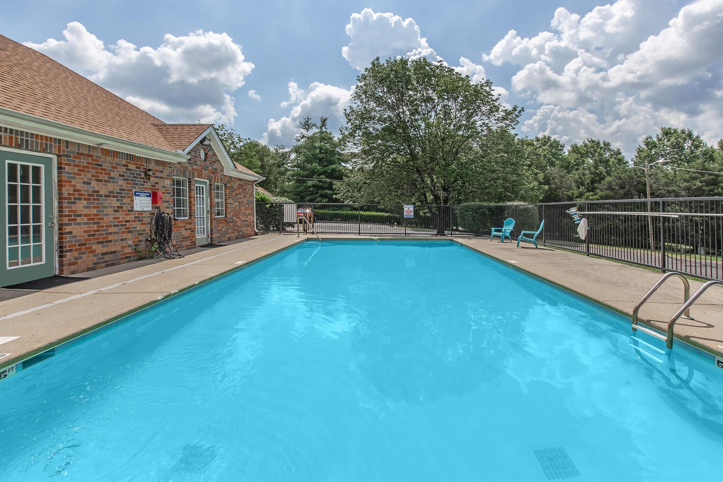 A clear blue swimming pool surrounded by a concrete deck. In the background, there is a brick building with large windows and a door, shaded by a tree. The sky is partly cloudy with puffy white clouds, and there are a couple of chairs near the pool. The scene is peaceful and inviting for relaxation.