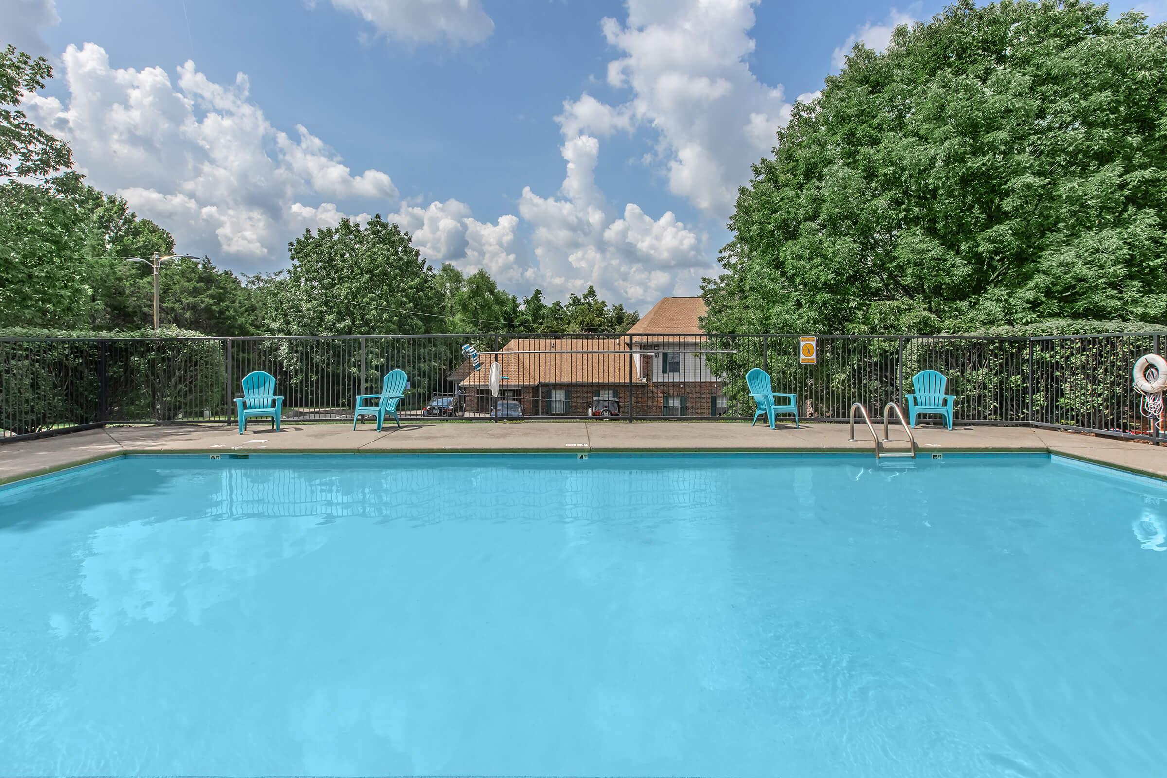 A clear blue swimming pool surrounded by a concrete deck, with several turquoise lounge chairs positioned nearby. Lush green trees provide shade in the background, and a brick building is visible beyond the pool area under a sky dotted with fluffy white clouds.