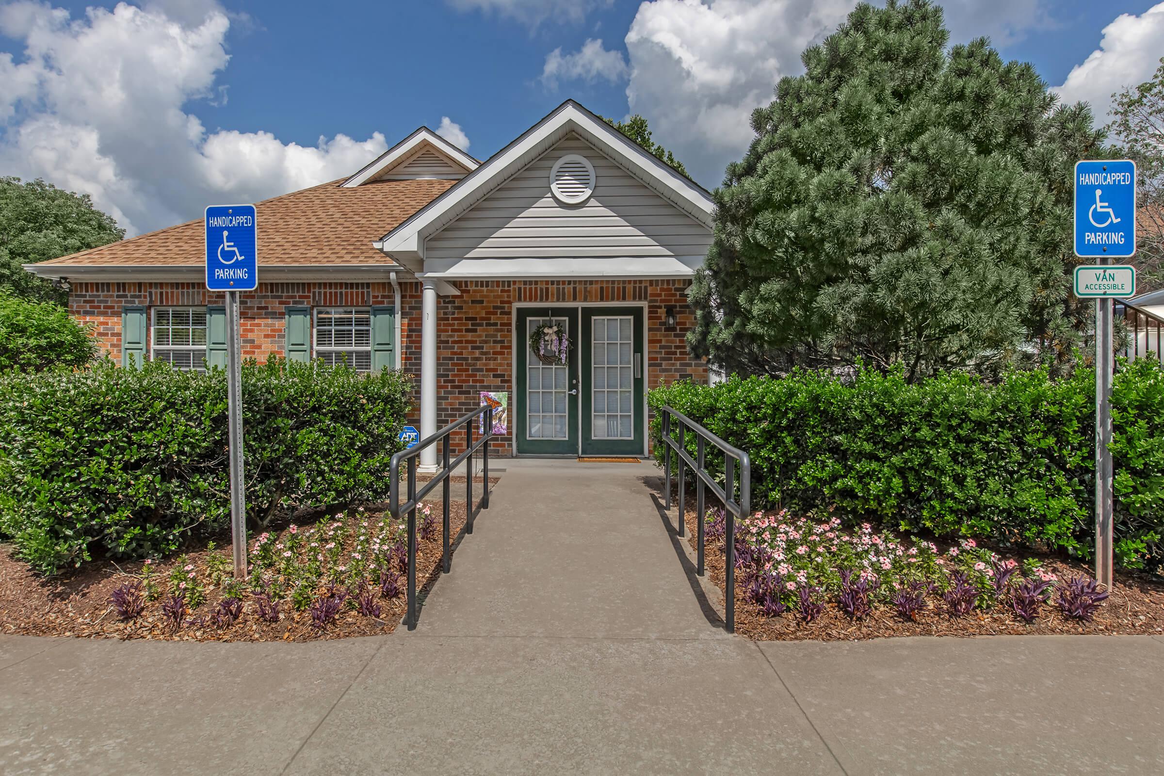 A single-story building with a porch and green doors, surrounded by well-maintained hedges and flower beds. Two blue signs indicate accessible parking, one on either side of a pathway leading to the entrance. The sky is partly cloudy, adding to the welcoming atmosphere.