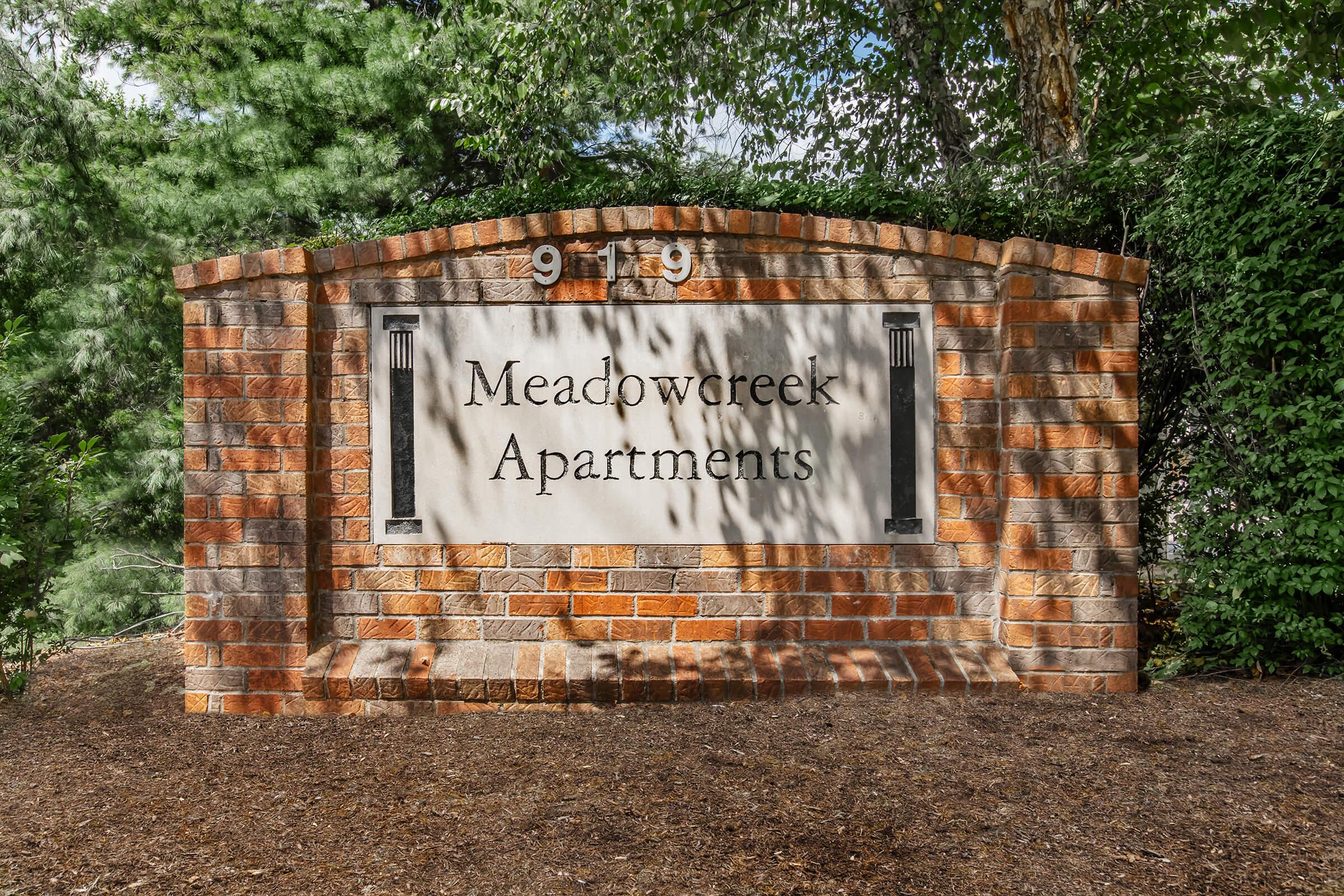 Stone sign for Meadowcreek Apartments with the number 919 displayed prominently. The sign features decorative elements and is surrounded by greenery, indicating a well-maintained residential area.