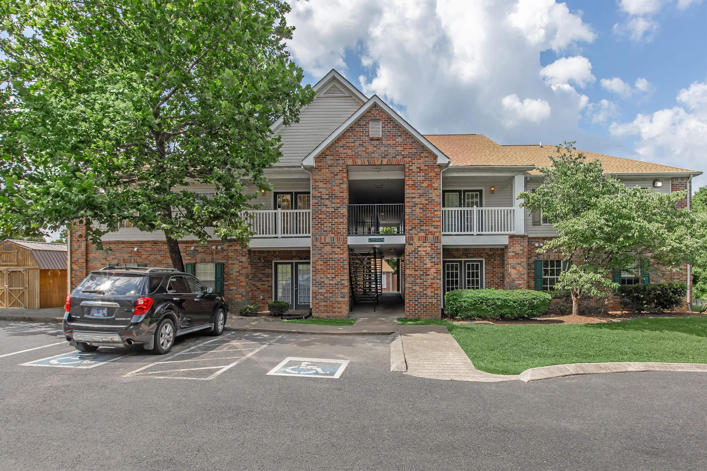 A two-story brick building with balconies, surrounded by green trees and landscaped lawns. A gray vehicle is parked in front, and a small parking area is visible. The sky is partly cloudy, creating a bright atmosphere.