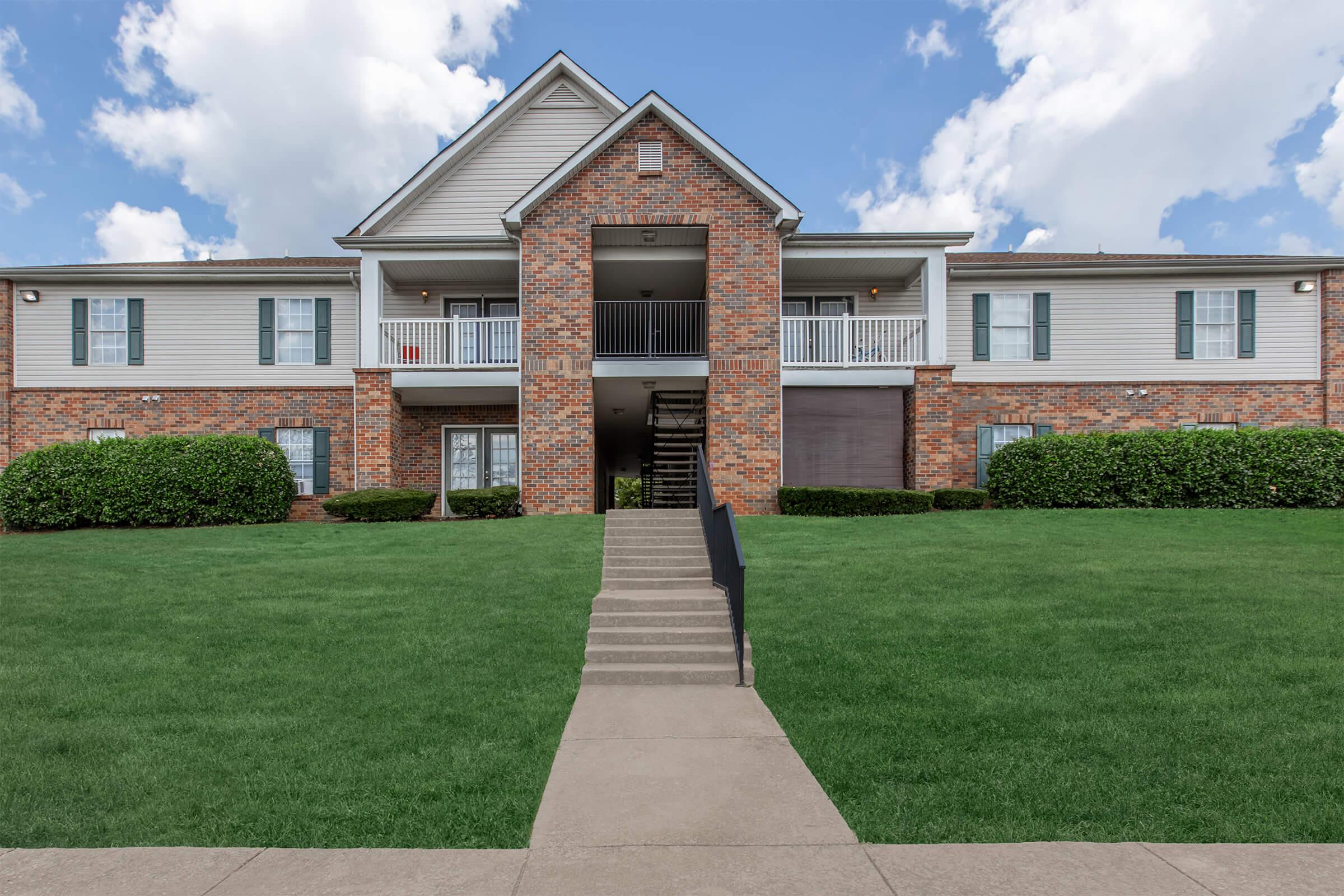 A brick apartment building with a central staircase leading to the upper level, flanked by green lawns and neatly trimmed bushes. The sky is partly cloudy, enhancing the bright appearance of the structure.