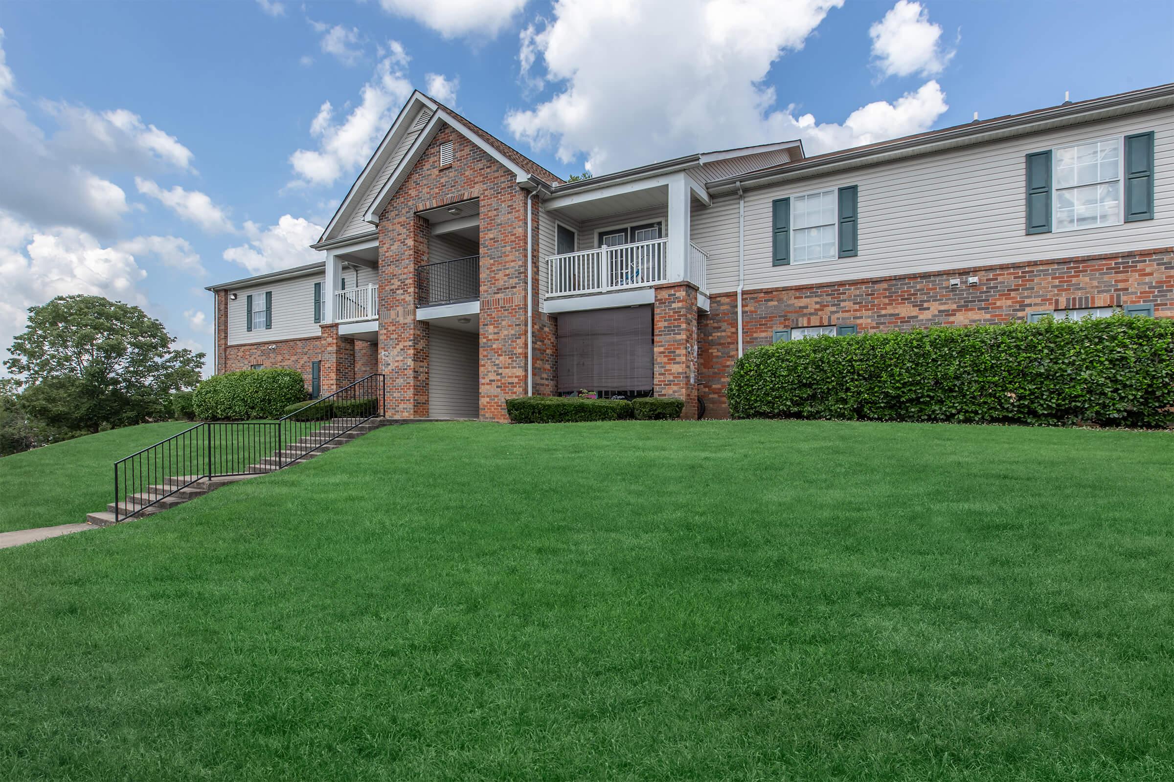 A brick apartment building with a landscaped lawn, featuring several balconies and windows. The sky is partly cloudy, and there are steps leading up to the entrance.