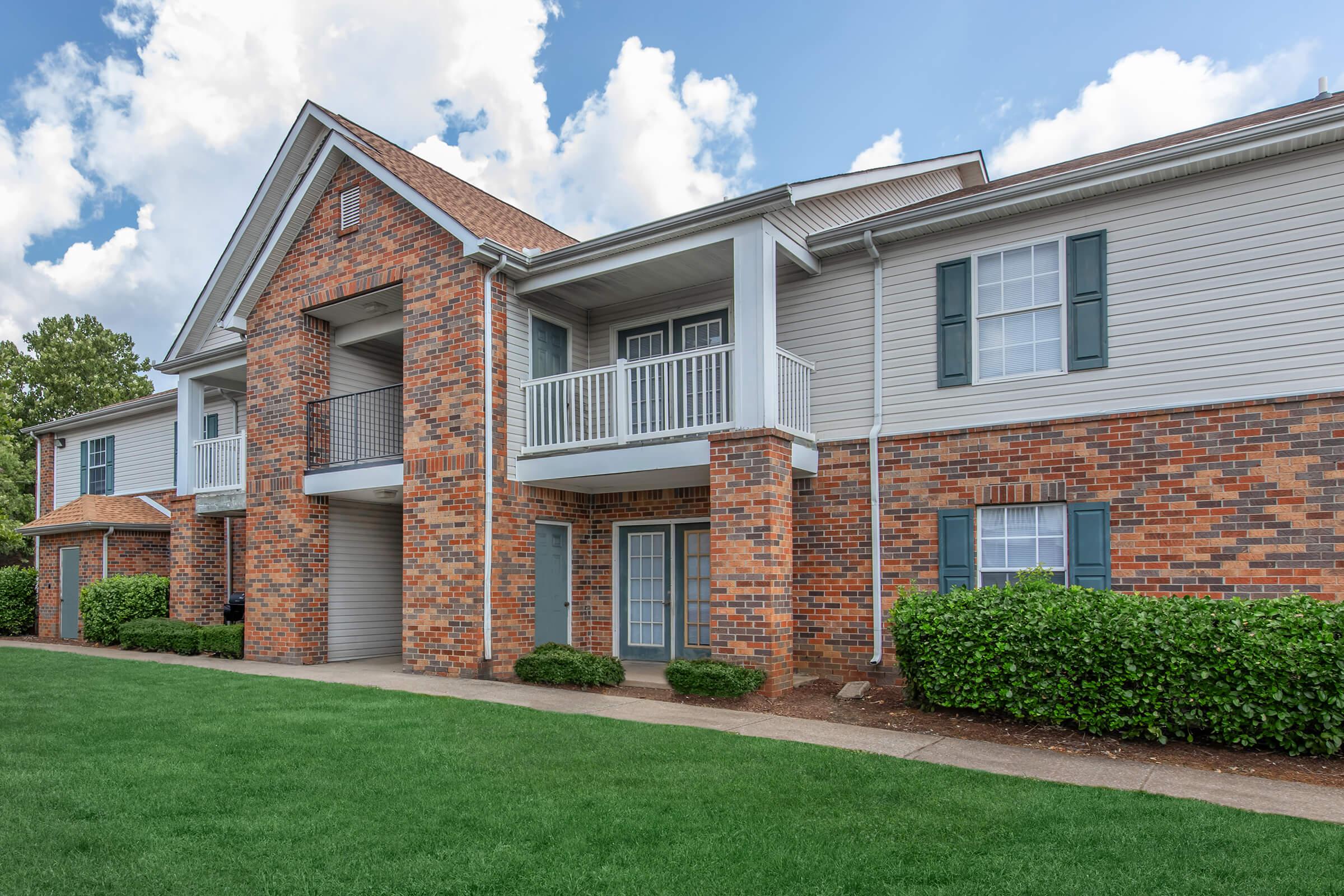 A well-maintained multi-unit residential building featuring a combination of brick and siding exterior. The building has balconies, green shrubbery, and a well-kept lawn. The sky is partly cloudy, contributing to a pleasant atmosphere.