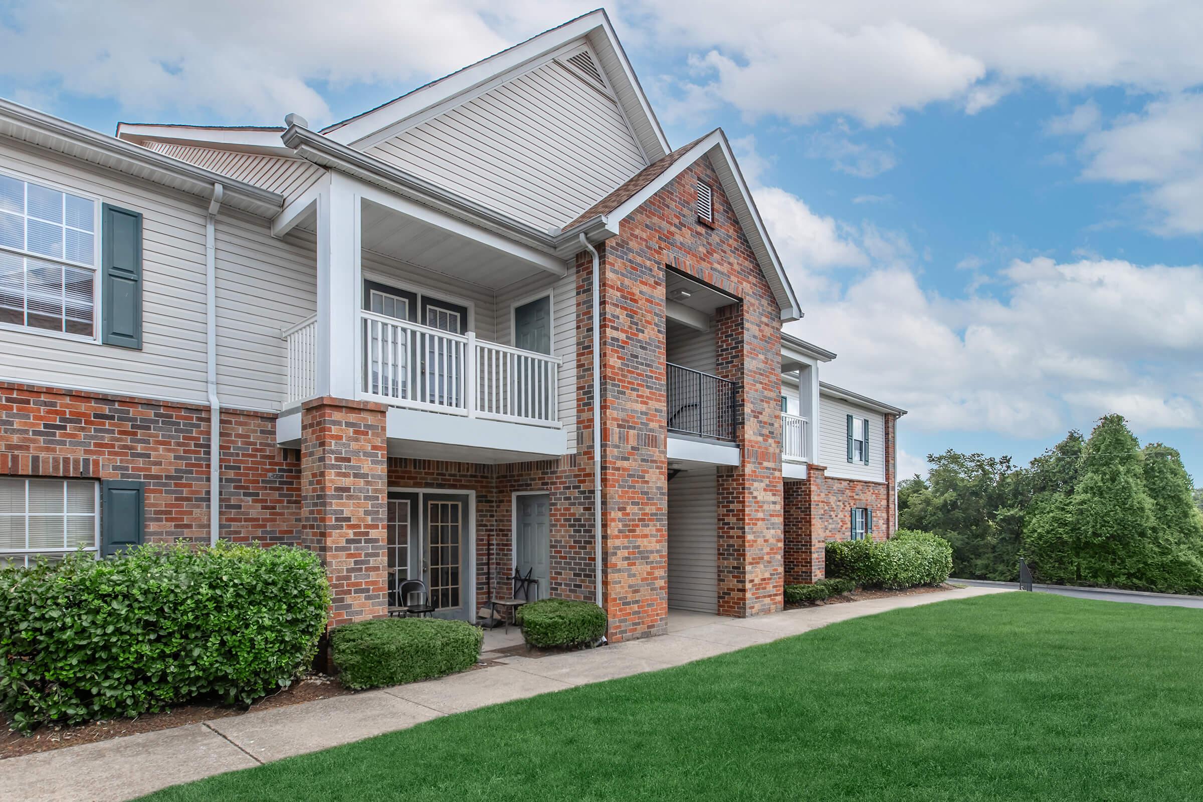 A charming apartment building featuring a brick facade and white siding. The structure has a covered porch with a black railing, flanked by well-maintained shrubs and a grassy lawn. The sky is partly cloudy, adding a fresh look to the residential setting.